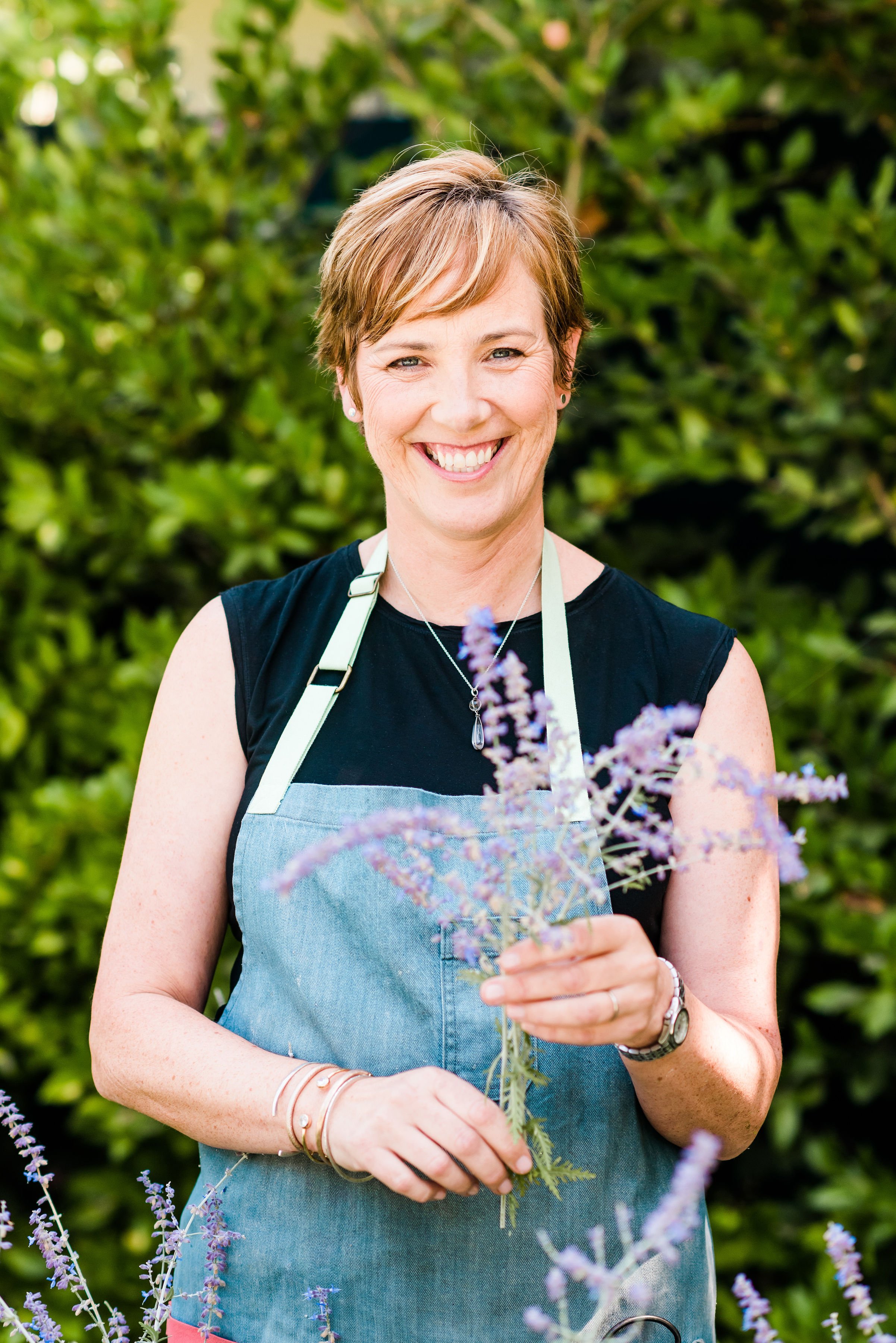 Smiling woman with short hair holding a bouquet of purple flowers outdoors, wearing a black sleeveless top and denim apron, surrounded by greenery.