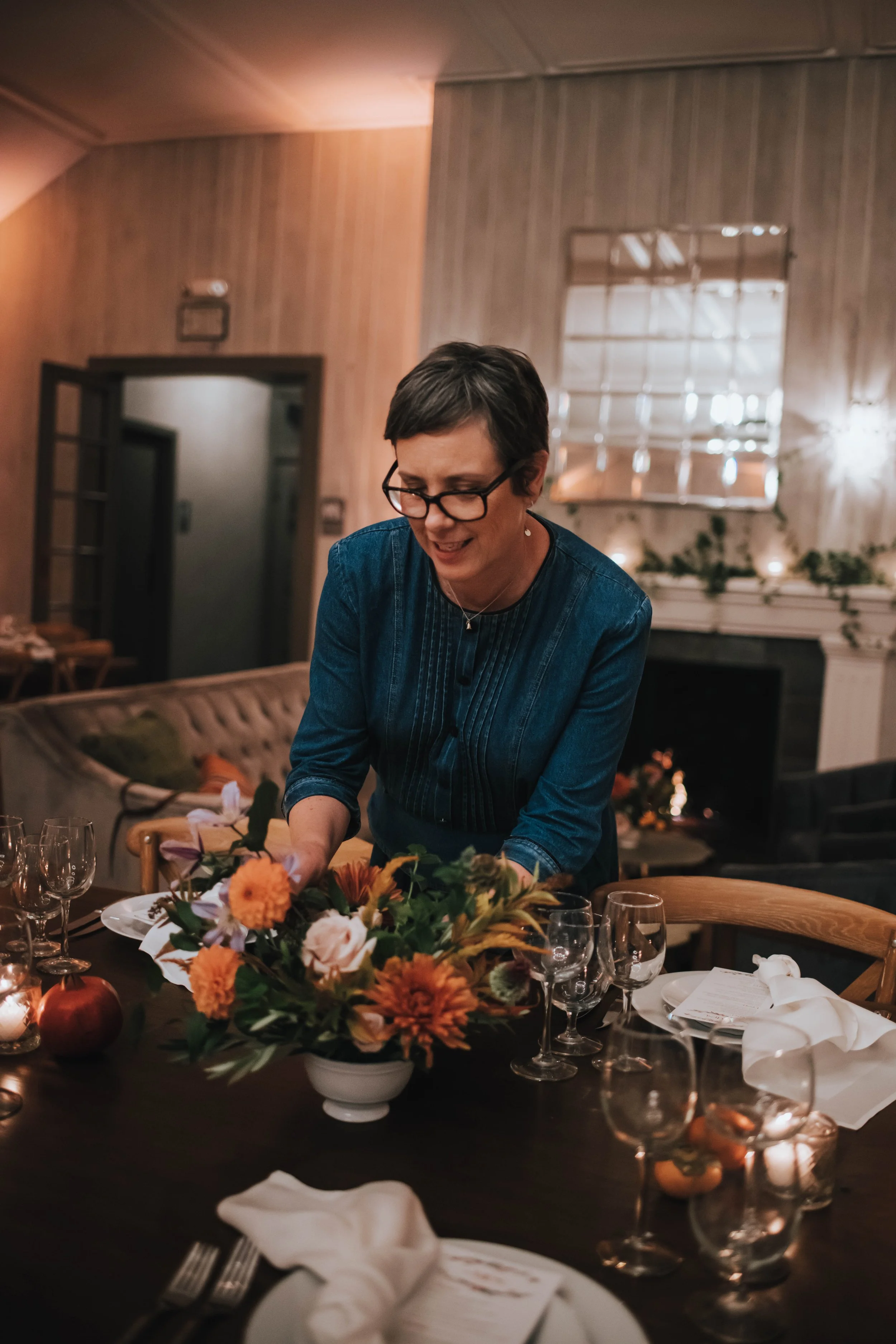 A woman with short dark hair, glasses, and a blue dress is standing at a dining table with a floral centerpiece, glasses, and place settings, in a warmly lit room with wood-paneled walls and a fireplace.