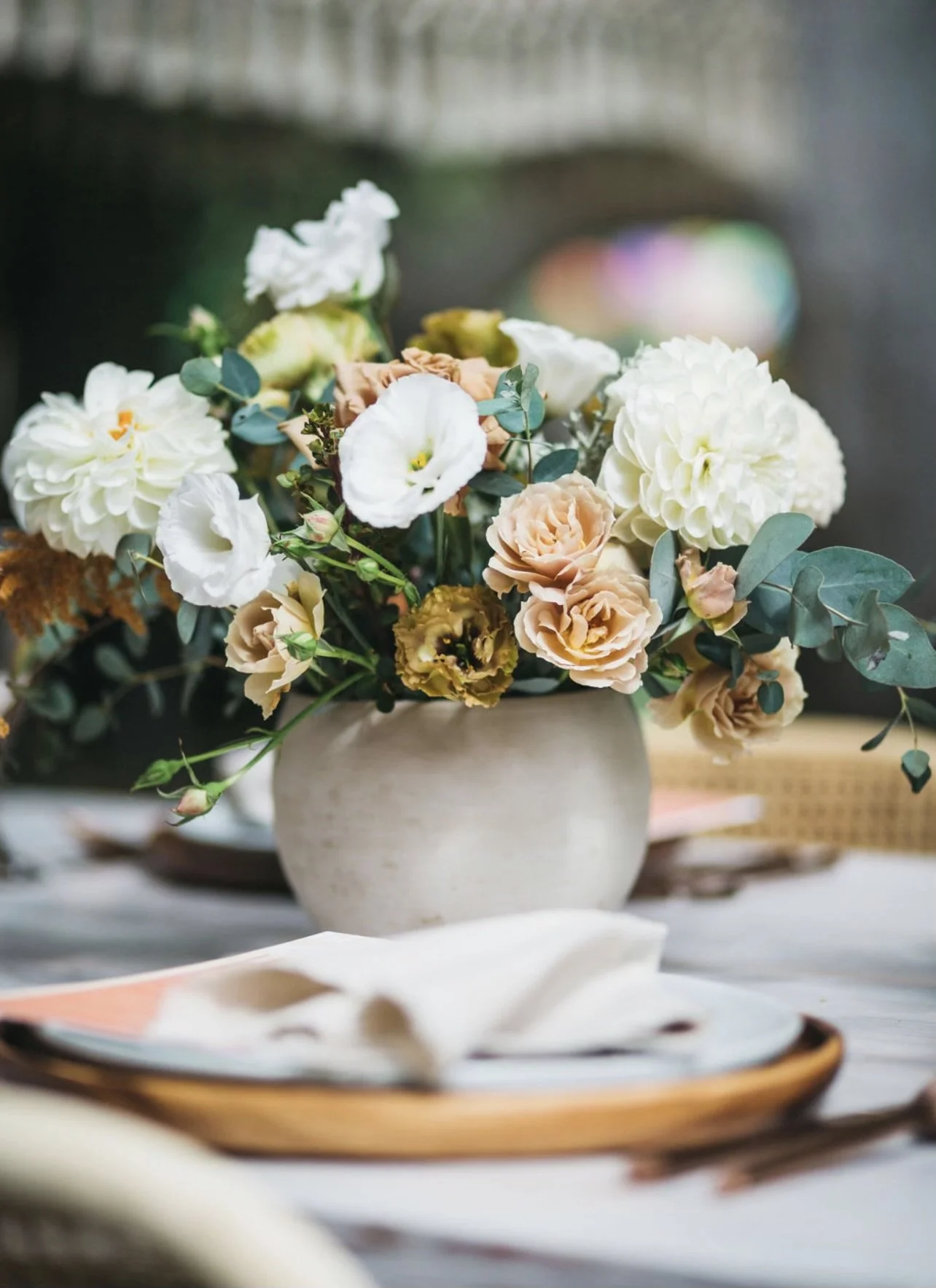 A bouquet of white, cream, and peach flowers arranged in a white ceramic vase on a table with a blurred background.