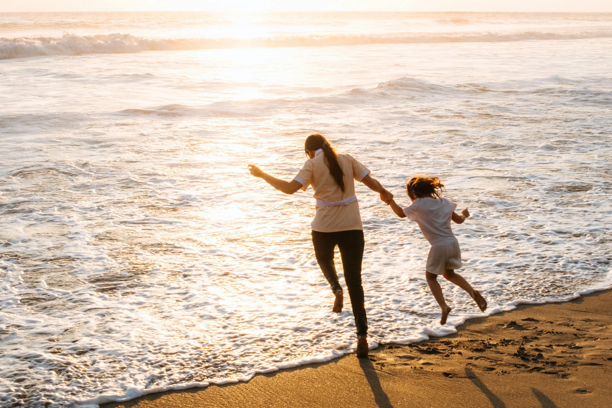 Adult and child playing by the ocean at sunset, holding hands and running through the shallow water with waves crashing on the sandy beach.