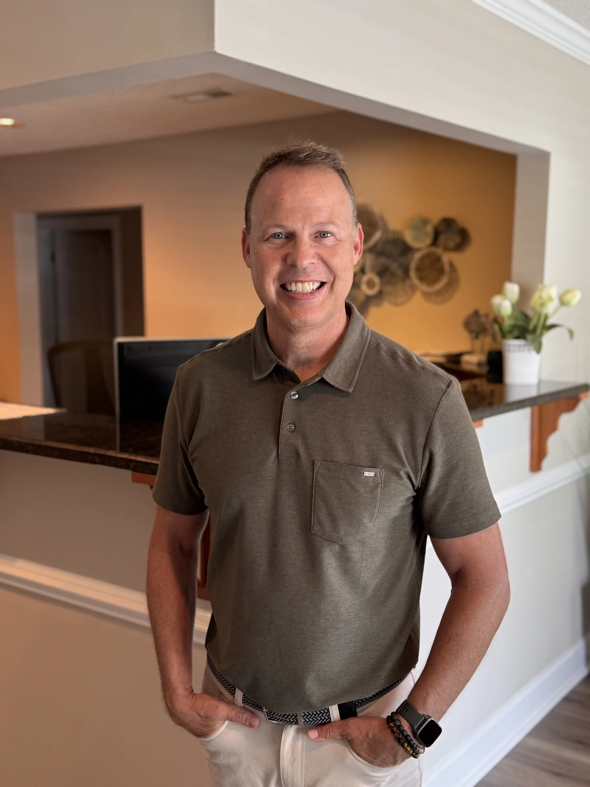 A smiling man standing with hands in his pockets indoors. He has short hair, wearing a brown polo shirt and white pants, with a dark bracelet and a smart watch on his left wrist. Background includes a decorative wall art and a vase with white flowers.