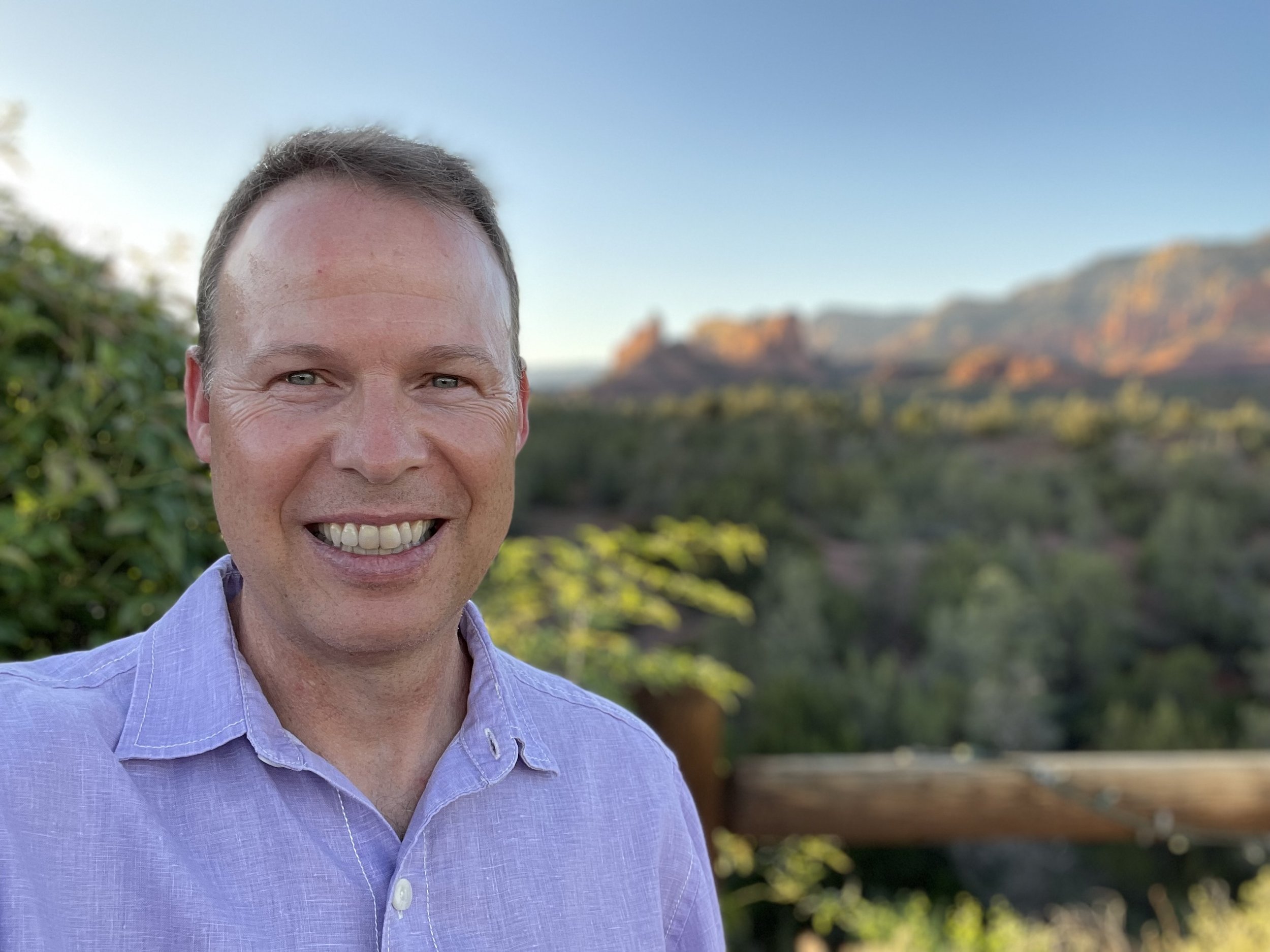 A smiling man with short brown hair wearing a lavender button-up shirt outdoors with desert mountains in the background.