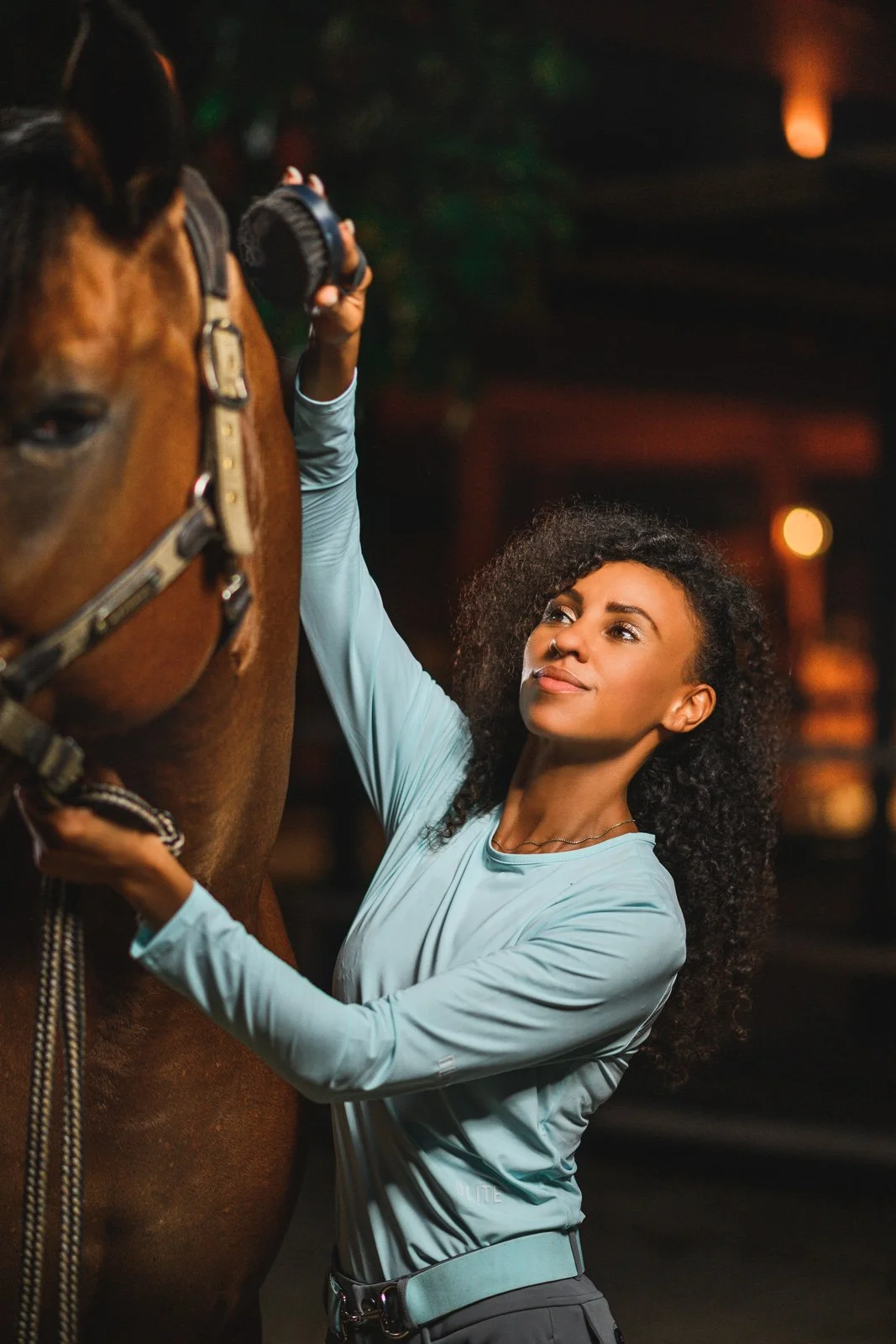 A woman with curly hair adjusting the strap of a brown horse's bridle indoors at night.