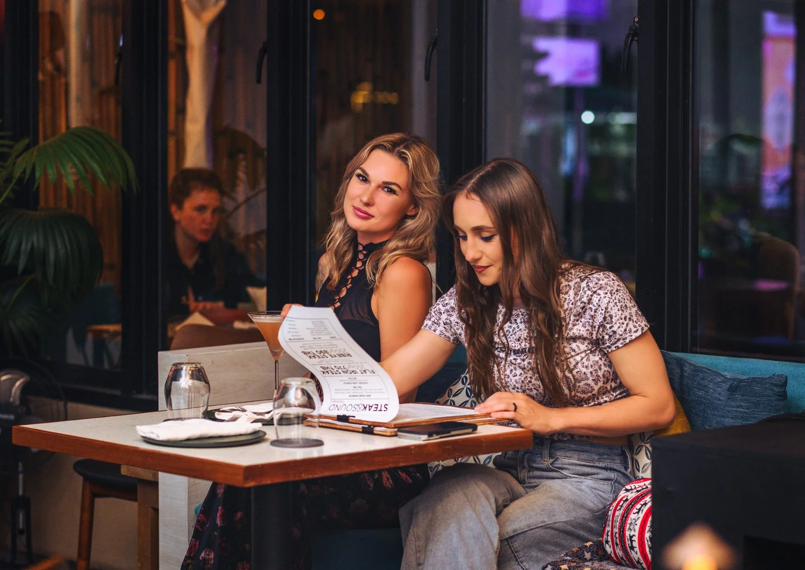 Two women sit at a restaurant table, one looking at a menu while the other looks at the camera. The table has wine glasses, a cocktail, and utensils. They are in a modern, cozy restaurant with large windows and indoor plants.