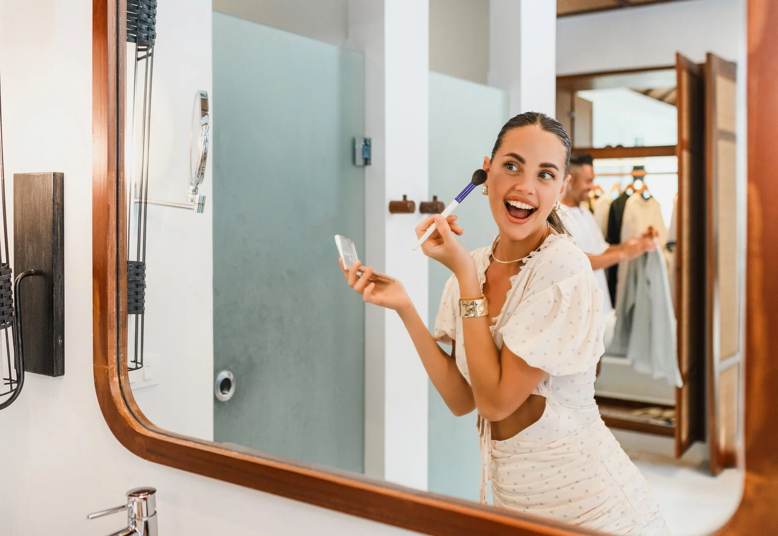 Woman applying makeup in front of a mirror at a clothing store, with a man in the background shopping.