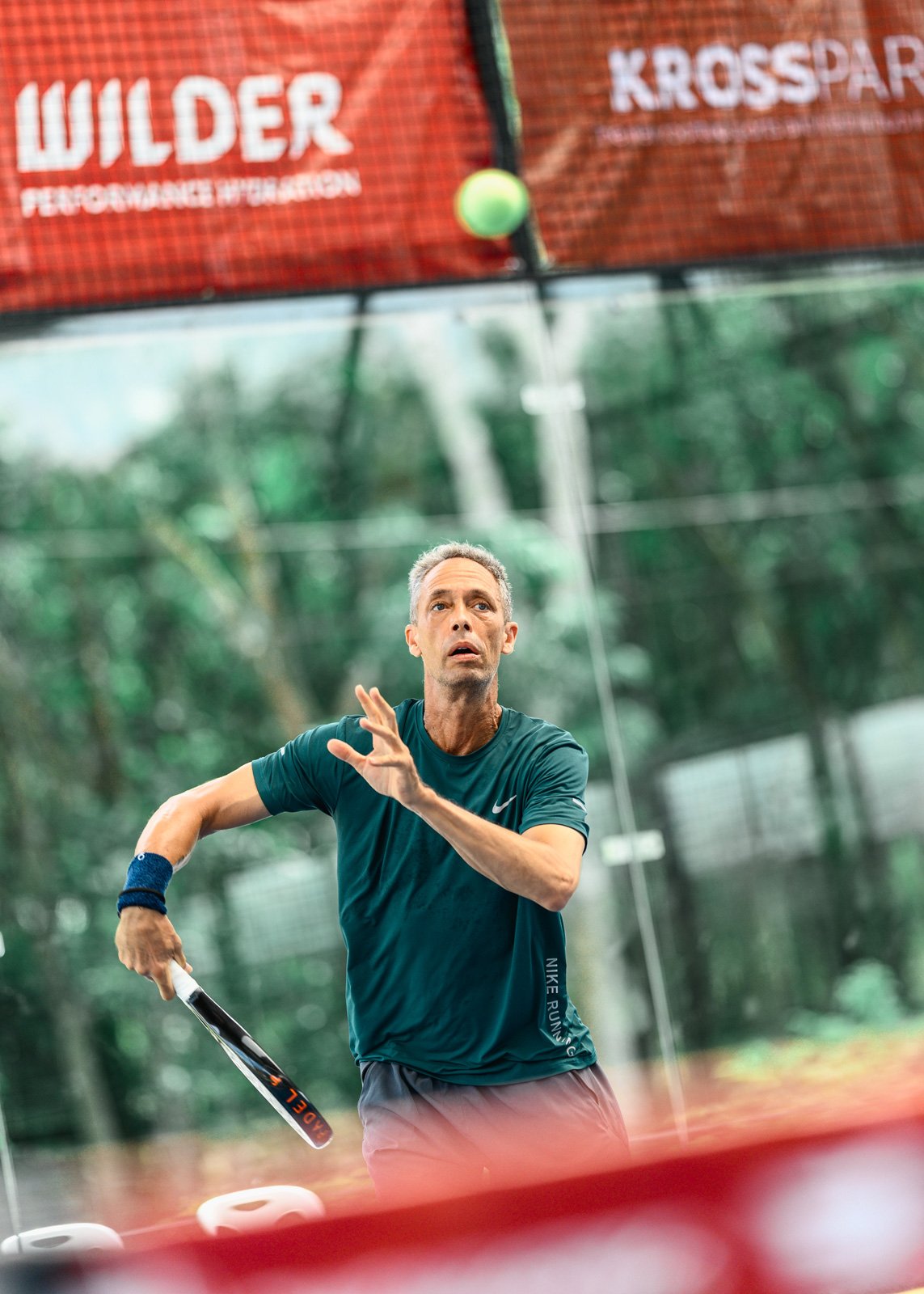 A man playing padel tennis outdoors near a net, with trees in the background.