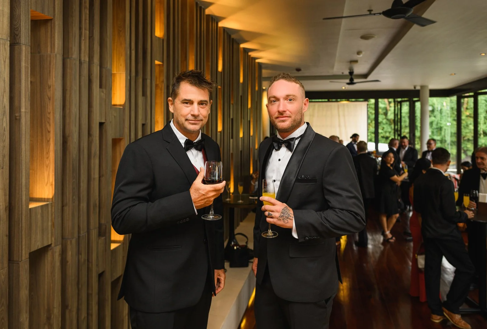 Two men in tuxedos holding glasses of wine and champagne at a formal event in a modern venue with wooden wall decor and large windows.