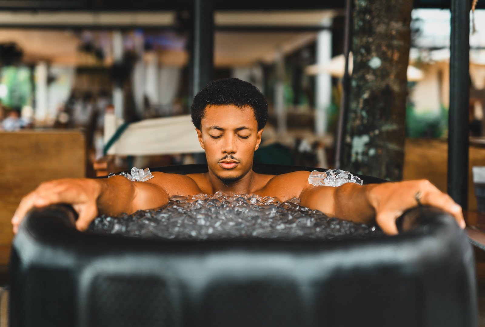 A man relaxes in a filled hot tub with closed eyes and outstretched arms in an outdoor spa setting.