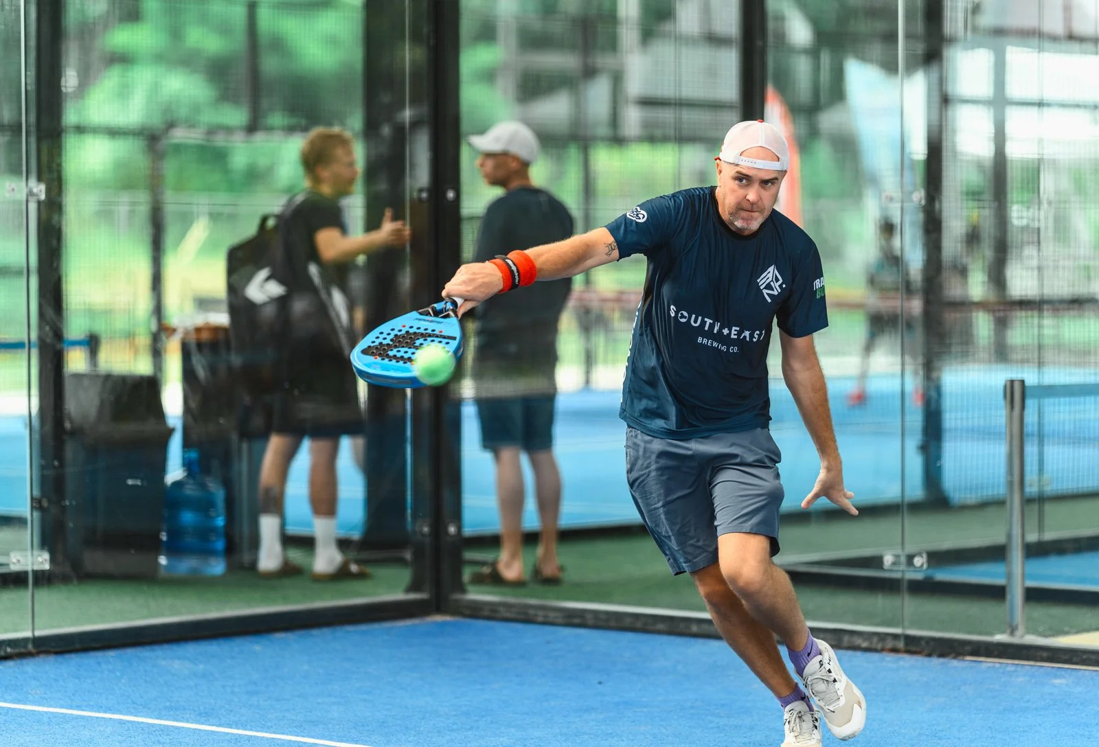 A man hitting a ball with a paddle on an indoor court, with people watching behind glass.