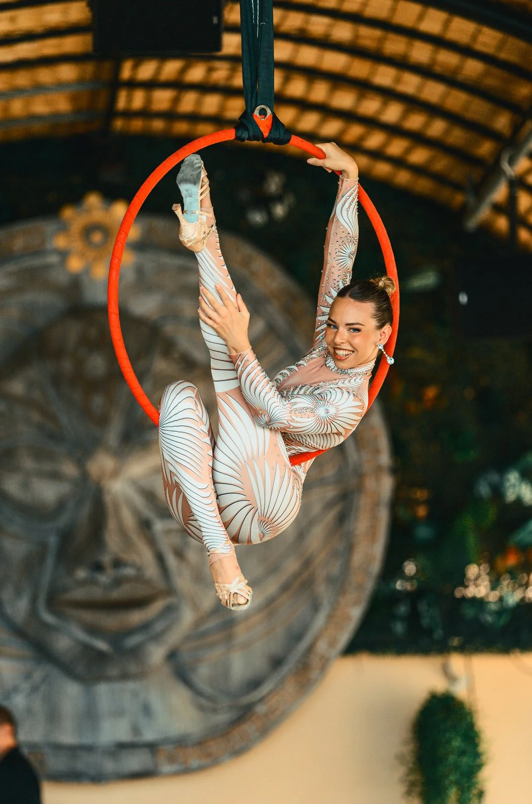 A female aerialist performing on a red aerial hoop, smiling and wearing a patterned jumpsuit with a floral design, in an indoor venue with a wooden ceiling and decorative sculpture in the background.