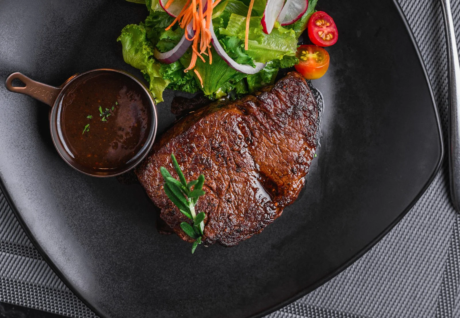 A plate with a cooked beef steak, fresh salad with cherry tomatoes, radish, shredded carrots, lettuce, and onion, and a small cup of dark sauce garnished with herbs.