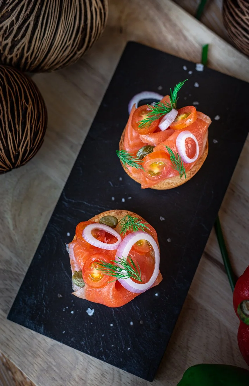 Two slices of smoked salmon appetizer on a black slate platter, topped with sliced onions, cherry tomatoes, capers, and garnished with dill and greenery.