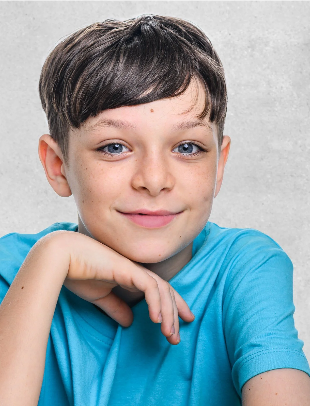 Close-up of a young boy with blue eyes and brown hair, wearing a light blue t-shirt, smiling and resting his chin on his hand against a plain grey background.