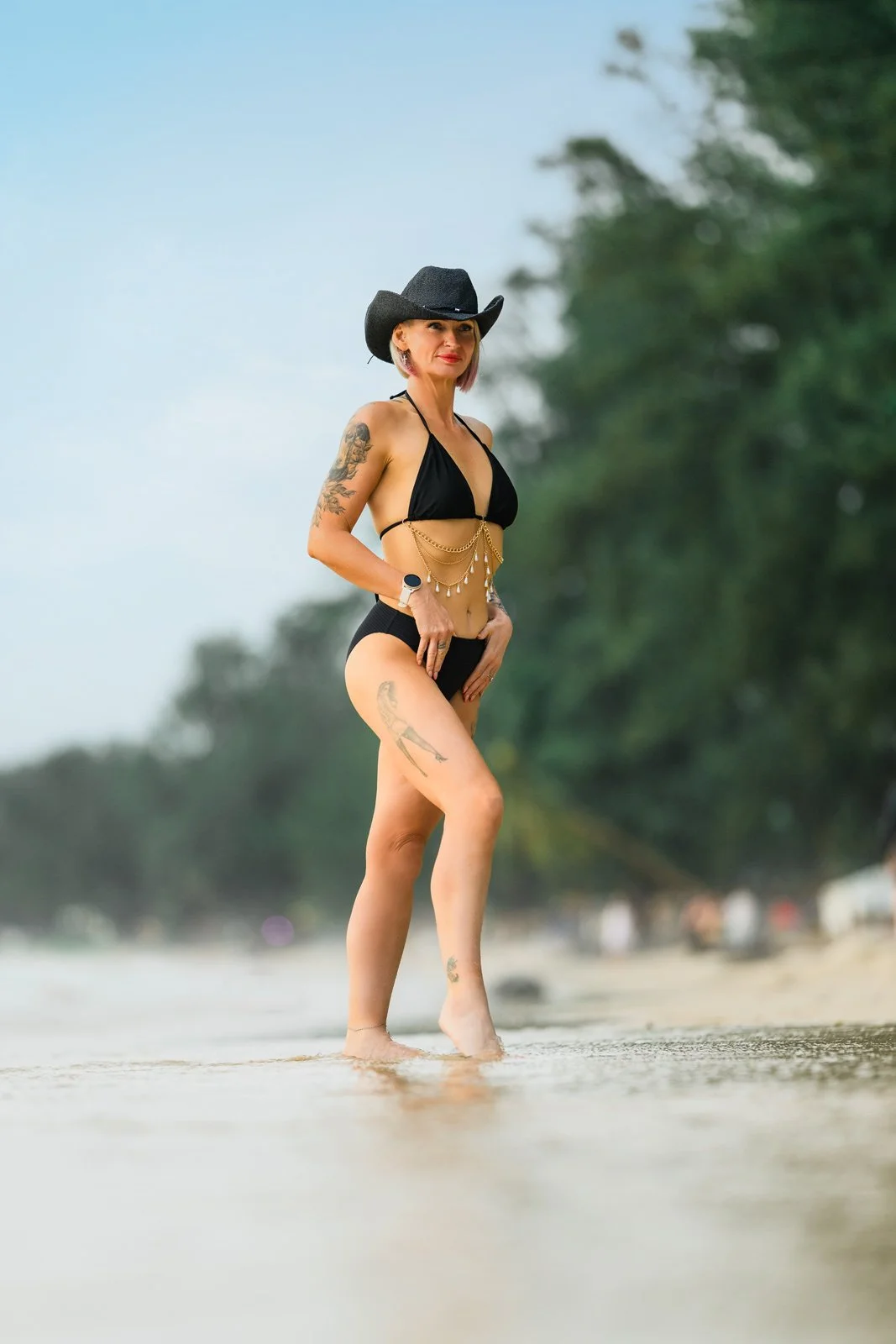 A woman standing on the beach wearing a black bikini, a black cowboy hat, tattoos on her arms and legs, and jewelry, with trees and the ocean in the background.