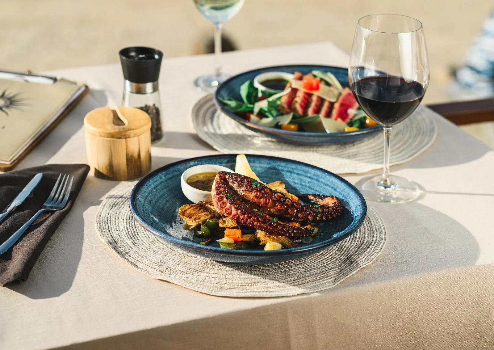 A table set with two blue plates of grilled octopus with vegetables and dipping sauce, two glasses of red wine, a salt grinder, a pepper shaker, a menu, and cutlery on a napkin, with sunlight shining on the table.