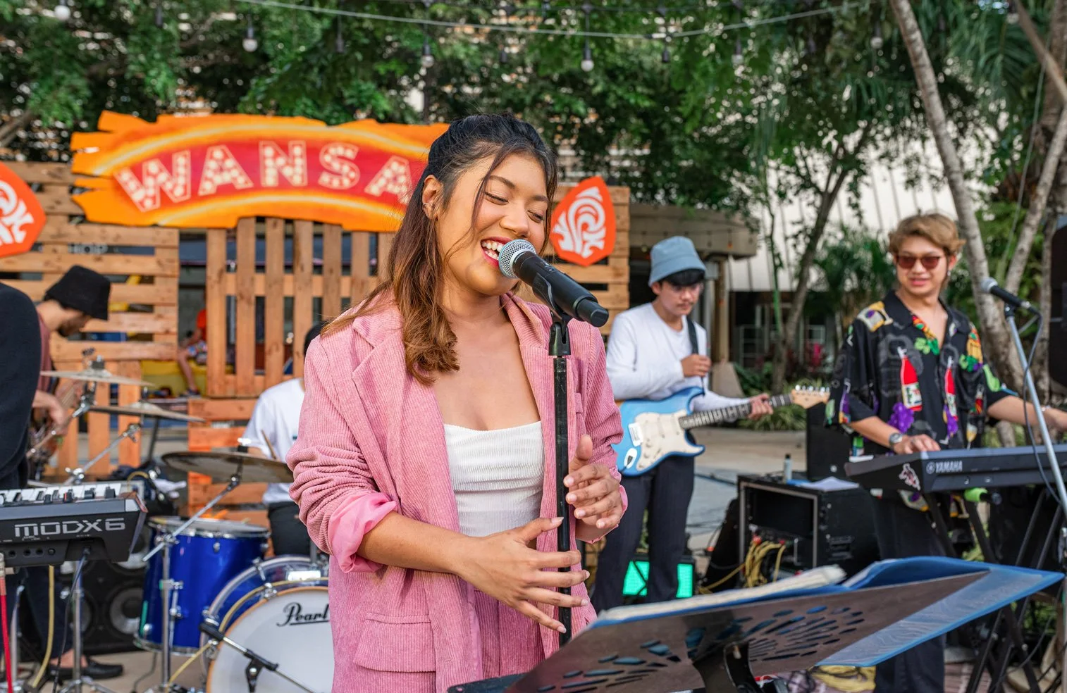 A woman singing and playing keyboard outdoors with a band in the background, under string lights and trees.
