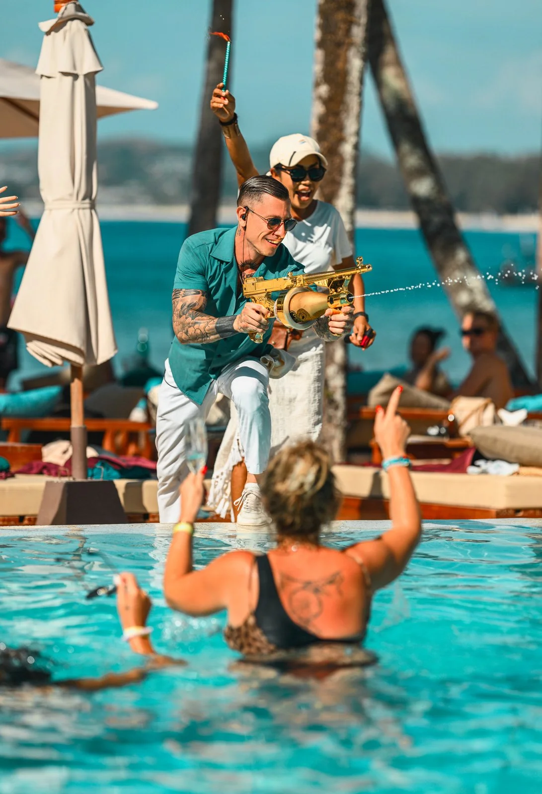 A person in a pool raising one arm, holding a sparking toy gun, while smiling. Another person near them is pointing at the person in the pool, and people in the background are relaxing near the water with palm trees visible.