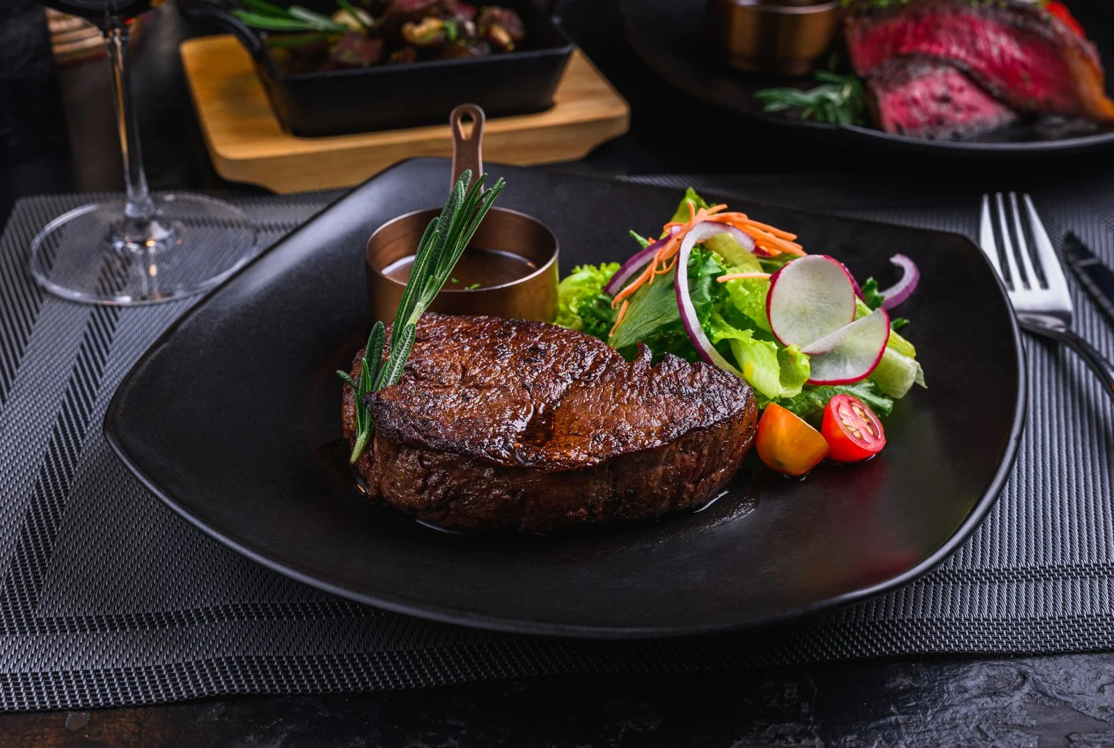 A plated meal featuring a grilled steak with a sprig of rosemary, accompanied by a mixed salad with cherry tomatoes, radish slices, shredded carrots, and lettuce, served with a small cup of sauce.