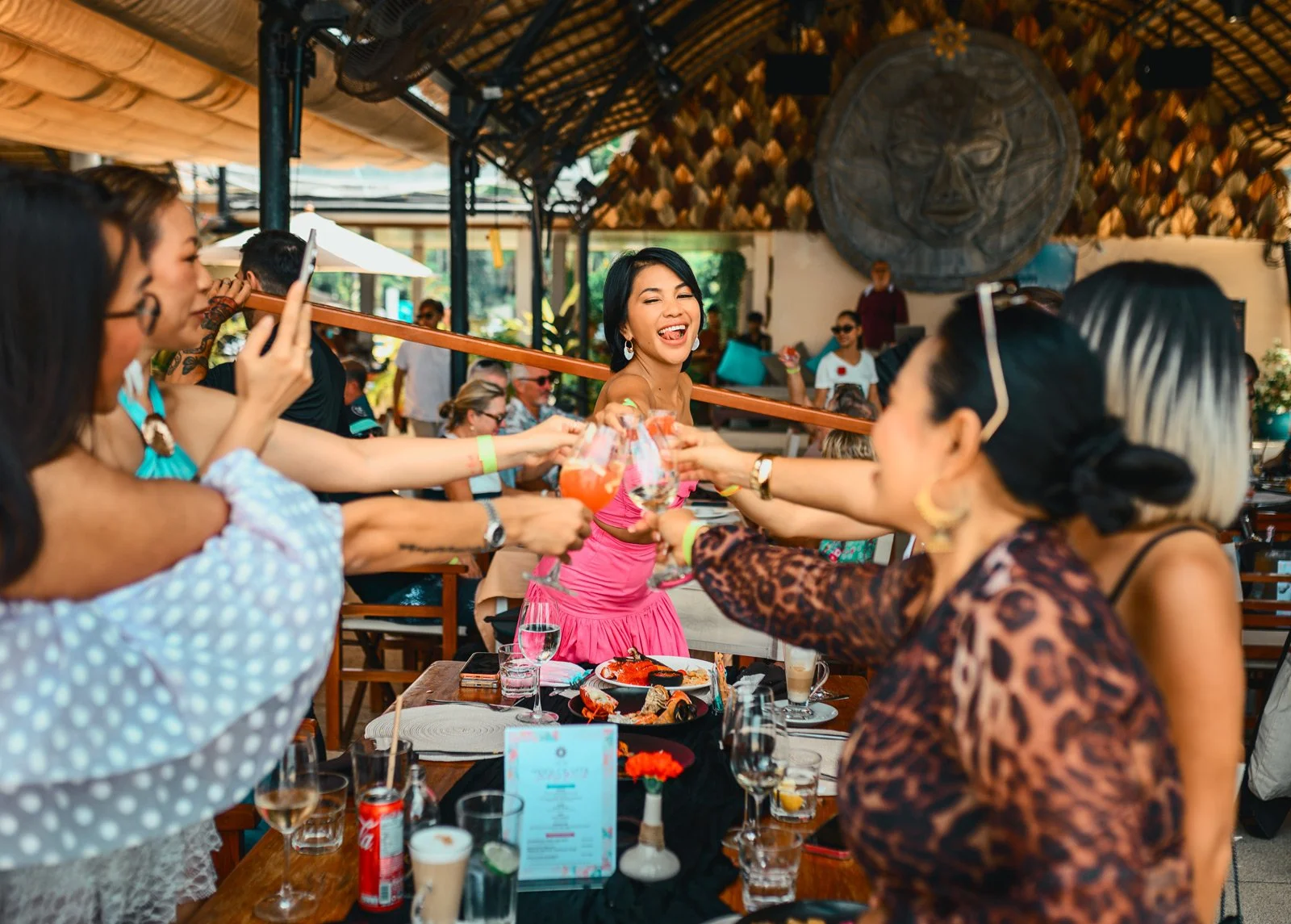 Group of women at a celebration table, with a woman in a pink dress smiling and laughing, as drinks are exchanged among friends in a lively restaurant or bar setting.