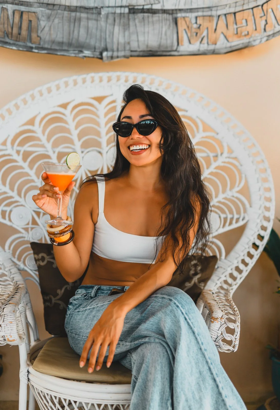 A woman with long dark hair wearing sunglasses, a white crop top, and light blue jeans sitting on a white wicker chair, holding a cocktail with a lime wedge, smiling broadly.