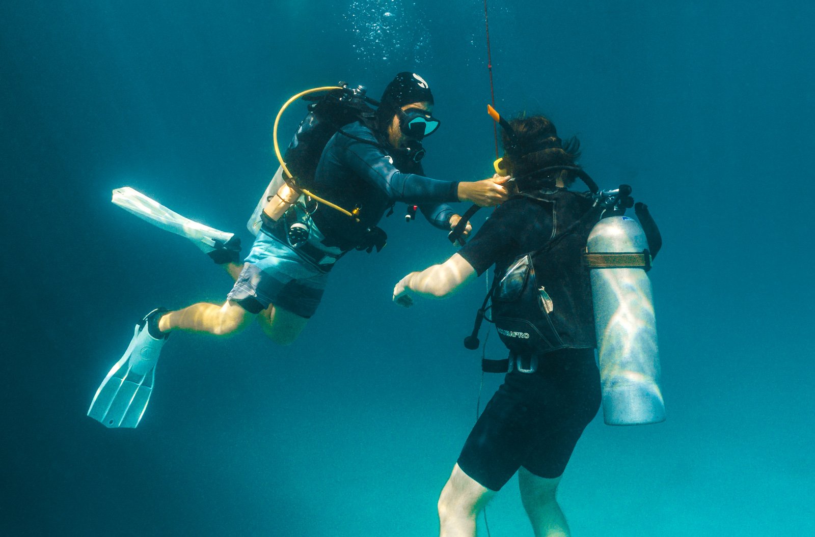 Two scuba divers underwater, one appears to assist the other with their diving gear.