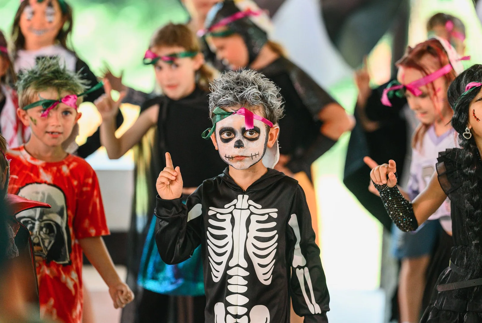 Children dressed in Halloween costumes, including a boy in a skeleton suit with face paint, wearing a headband, and others in various costumes with face paint and accessories, participating in a costume party or celebration.