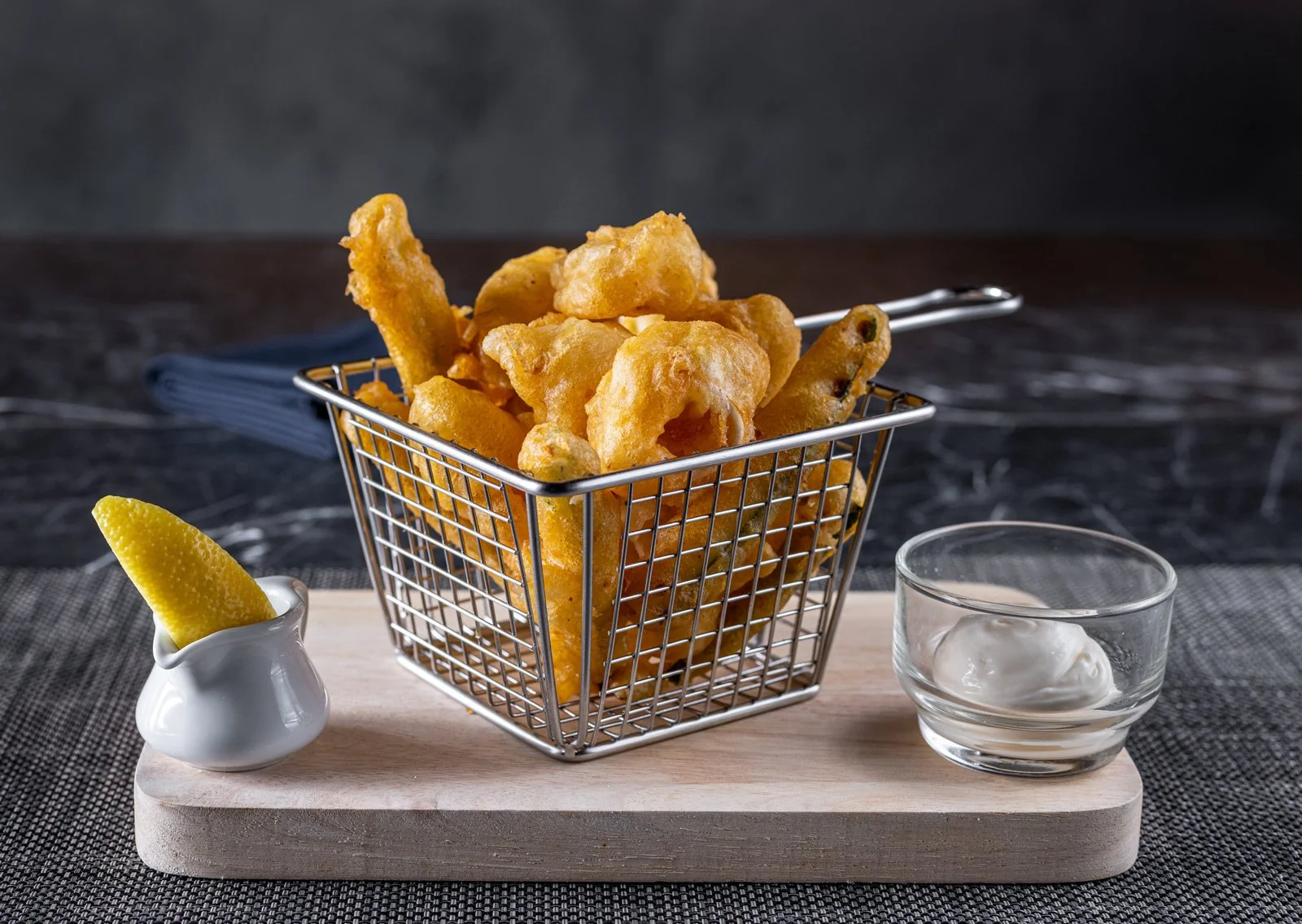 A wire basket filled with fried fish fillets and chips on a wooden board, with a lemon wedge in a small white pitcher and a small glass bowl of tartar sauce, all on a dark, textured surface.