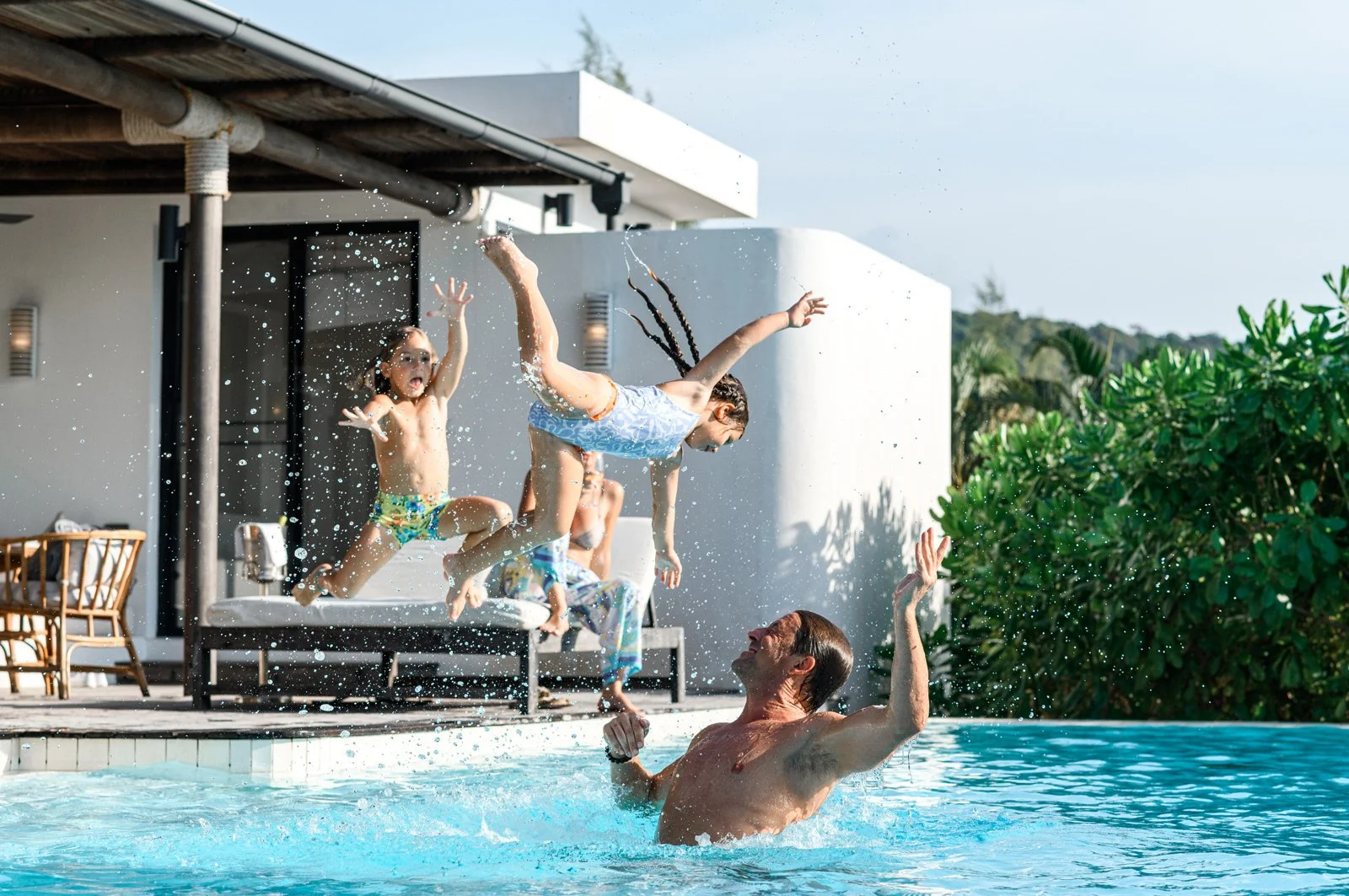 A man is in a swimming pool throwing a child into the air, while another child on a trampoline nearby reacts excitedly, with water splashing around on a poolside backyard with greenery and a modern house in the background.