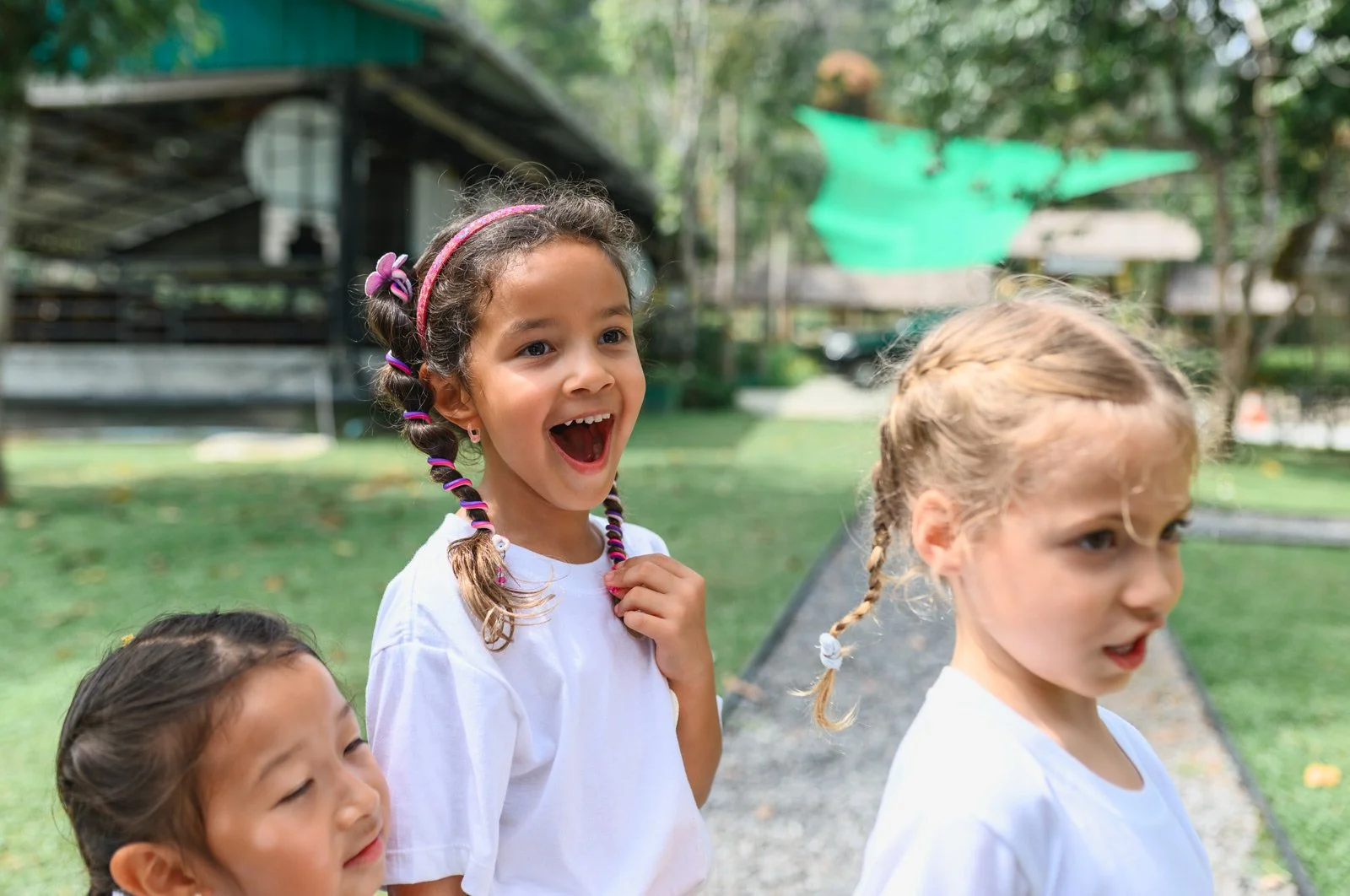 Three young girls outdoors, one girl smiling with colorful pigtails, another girl with blonde hair looking away, and a third girl with dark hair, all wearing white shirts, in a green park setting.