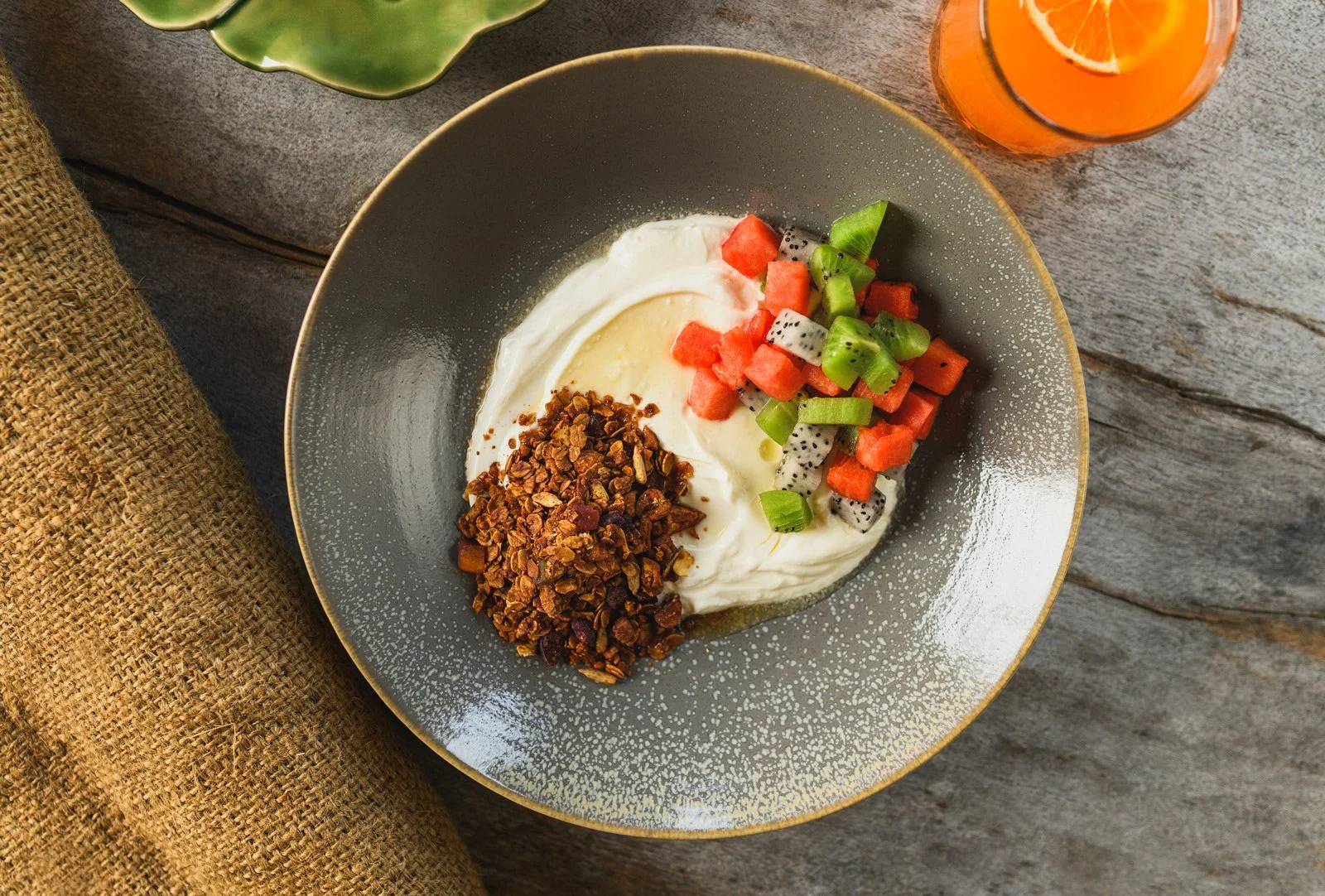 A bowl containing yogurt topped with granola and mixed fruits such as watermelon, kiwi, and dragon fruit, with a glass of orange juice and a green leaf on a wooden table.