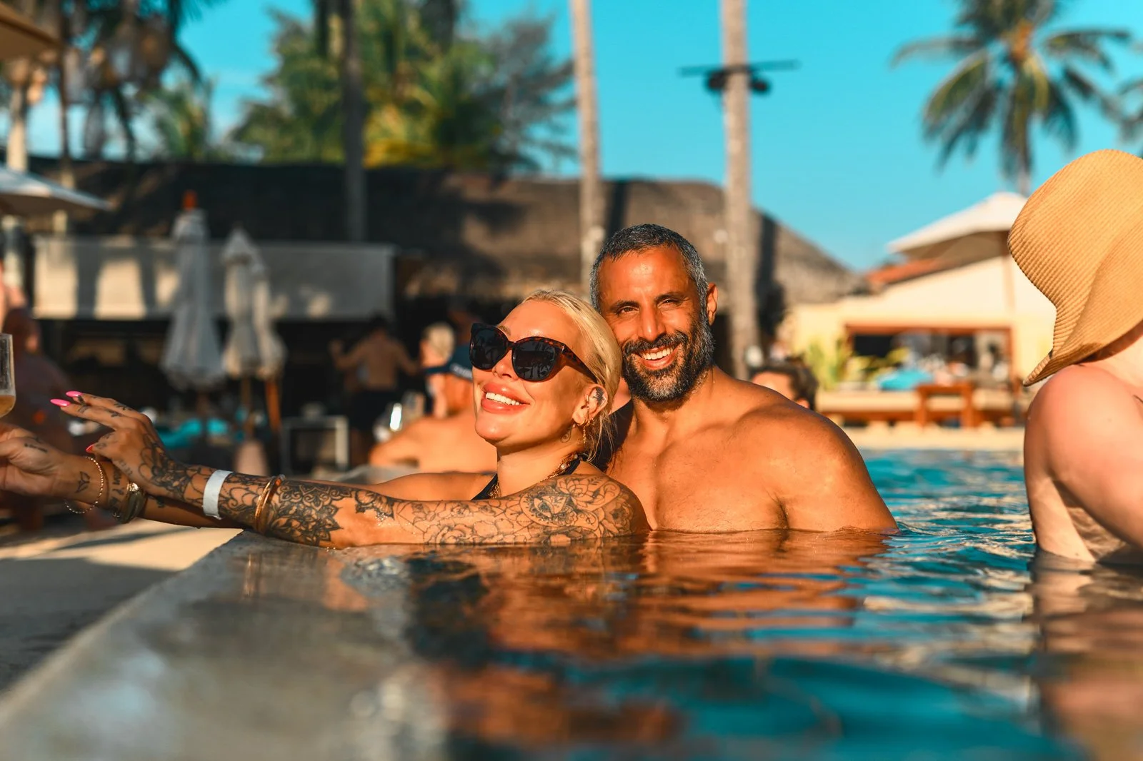 A smiling couple enjoying time in a swimming pool at a tropical resort, with palm trees and other people in the background.