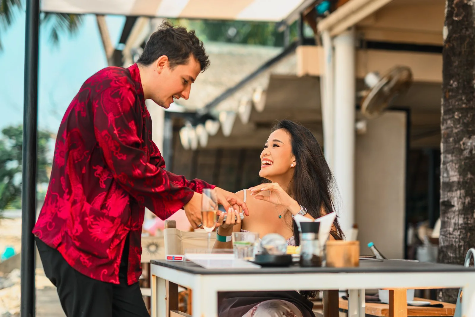 A man in a red patterned shirt is serving a drink to a woman sitting at a table outdoors. The woman is smiling and wearing a white sleeveless top. The table has various items including a camera, a glass, and a plate, and the setting appears to be a c