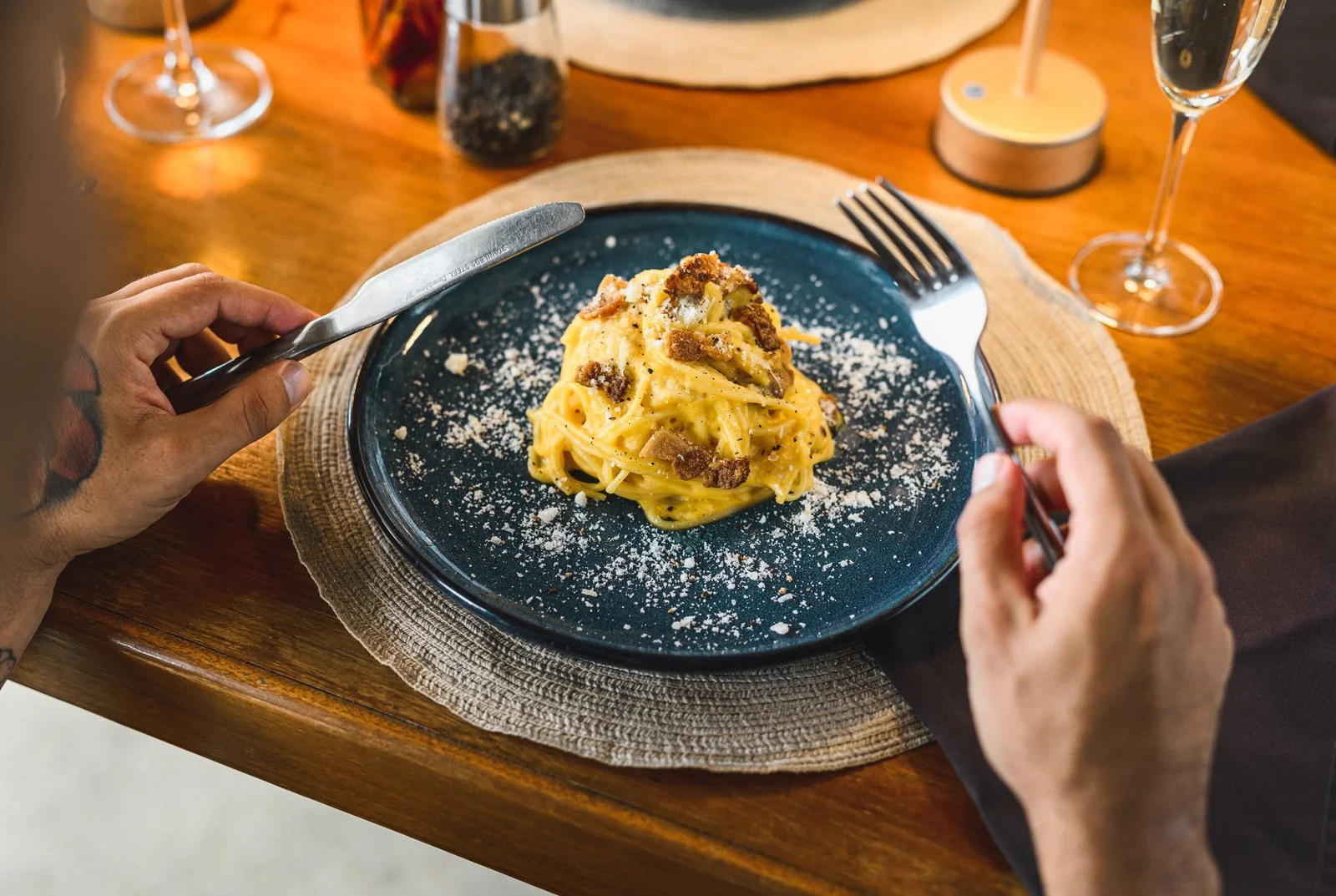 Person cutting into a plate of creamy pasta with black pepper and grated cheese, on a wooden table with wine glasses and condiments in the background.