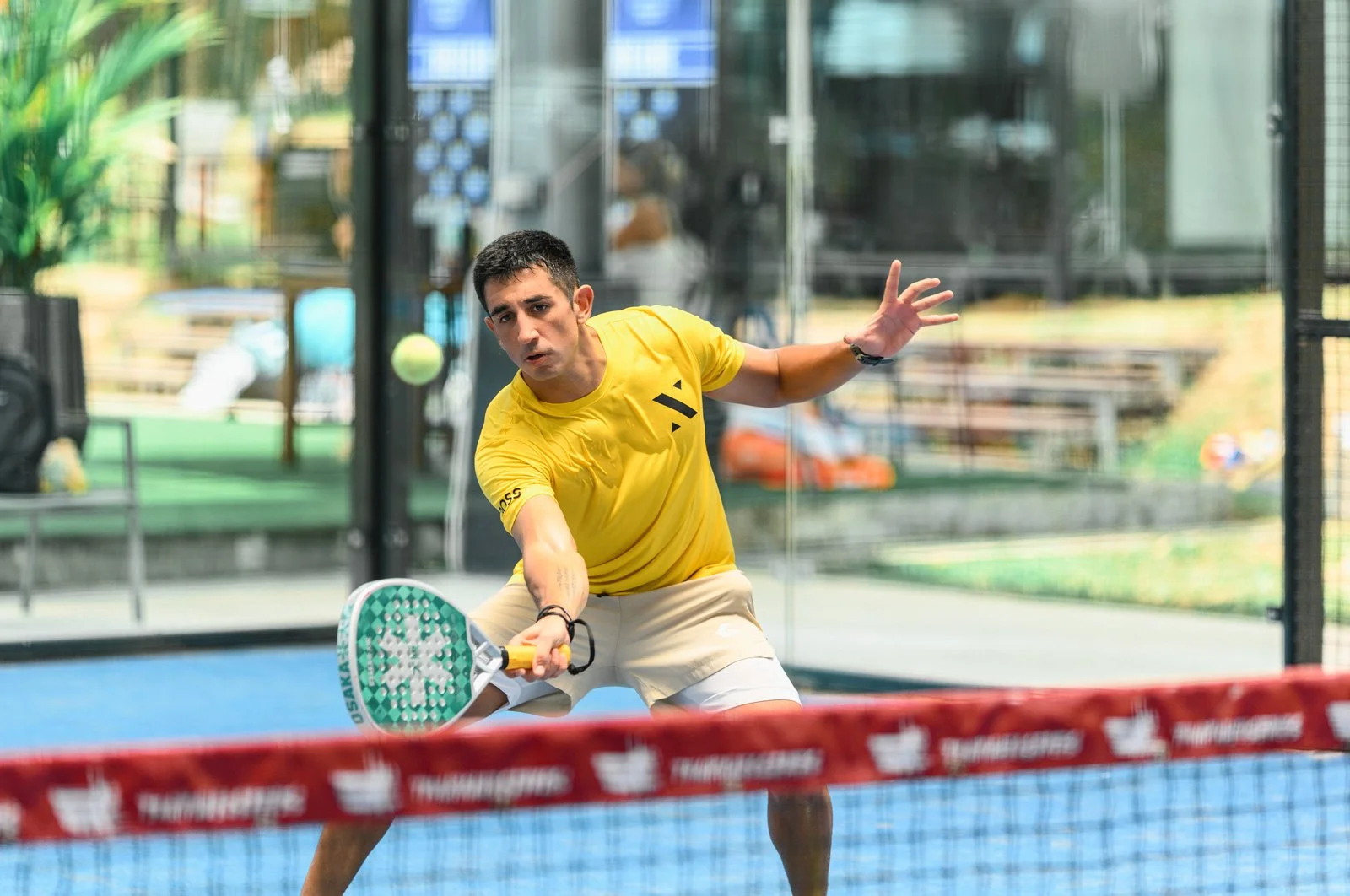Man playing padel on an Padel Phuket court, wearing a yellow shirt and beige shorts, holding a paddle, preparing to hit the ball.
