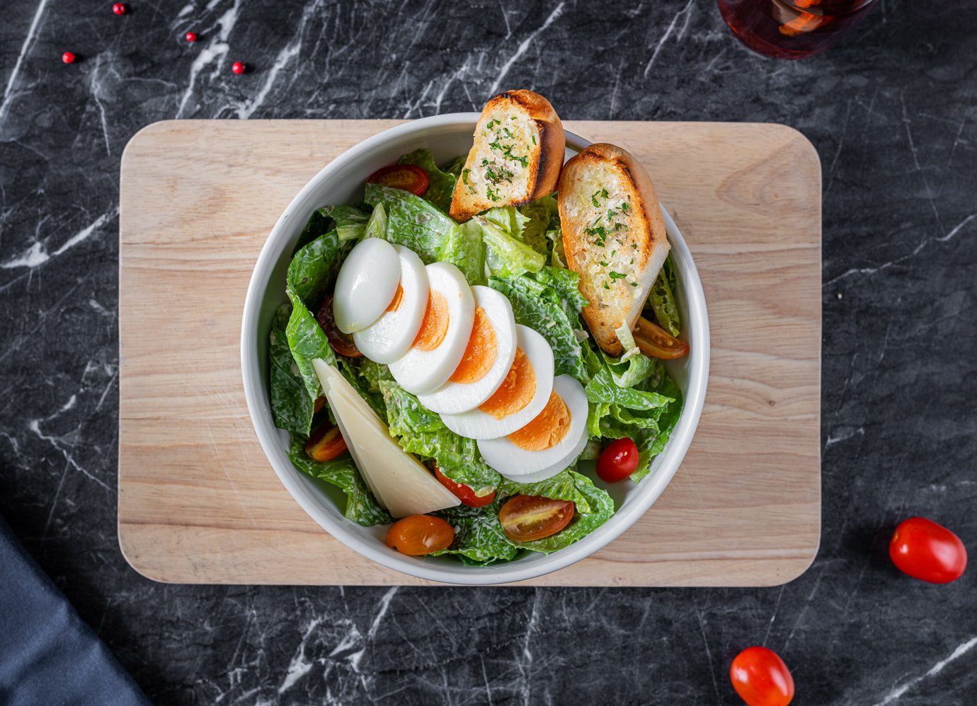 A bowl of salad with sliced hard-boiled eggs, cherry tomatoes, lettuce, and toasted bread slices on a wooden cutting board, on a dark marble surface.