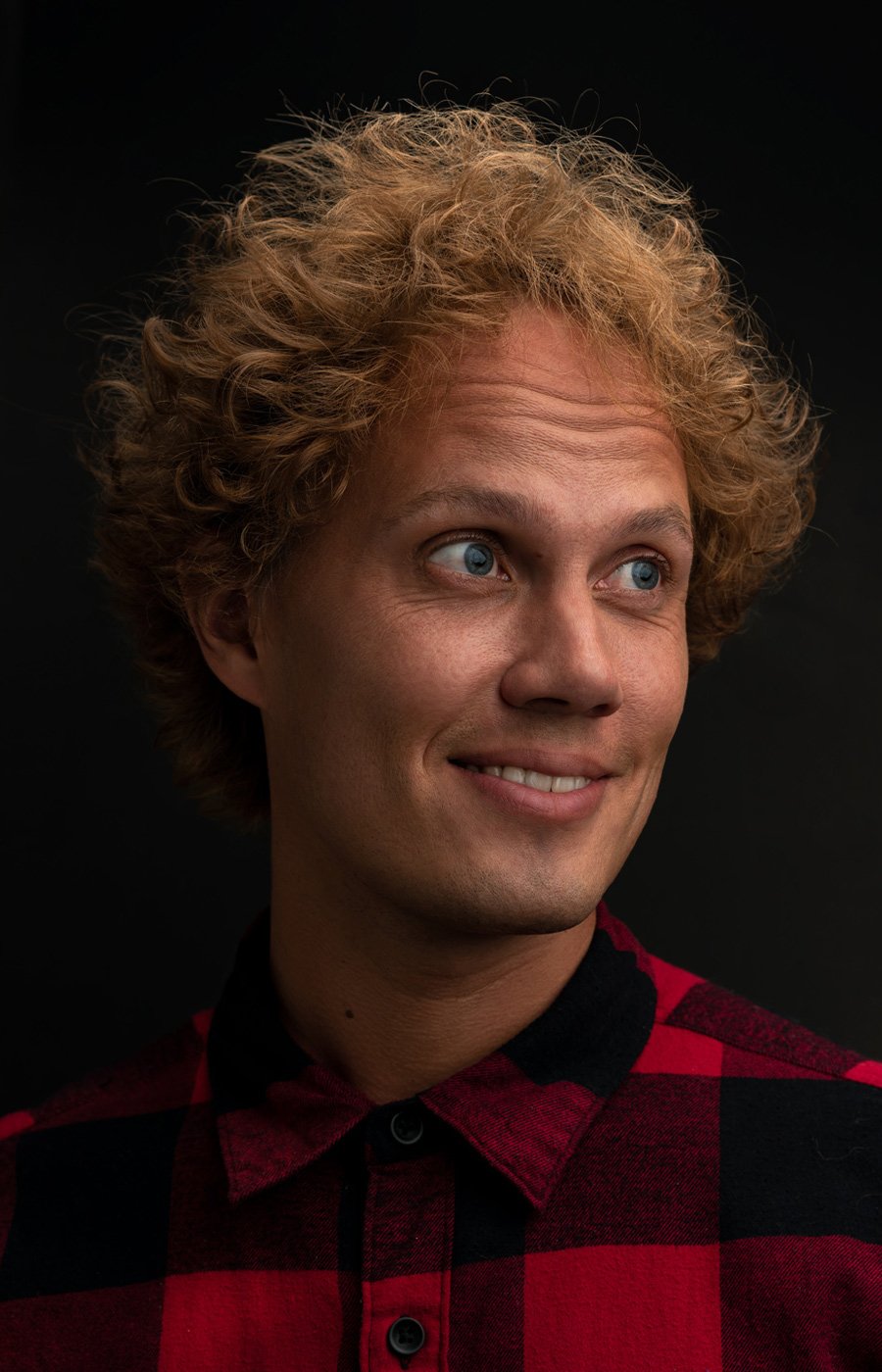 Close-up portrait of a young man with curly blonde hair and blue eyes, smiling, wearing a red and black checkered shirt against a black background.
