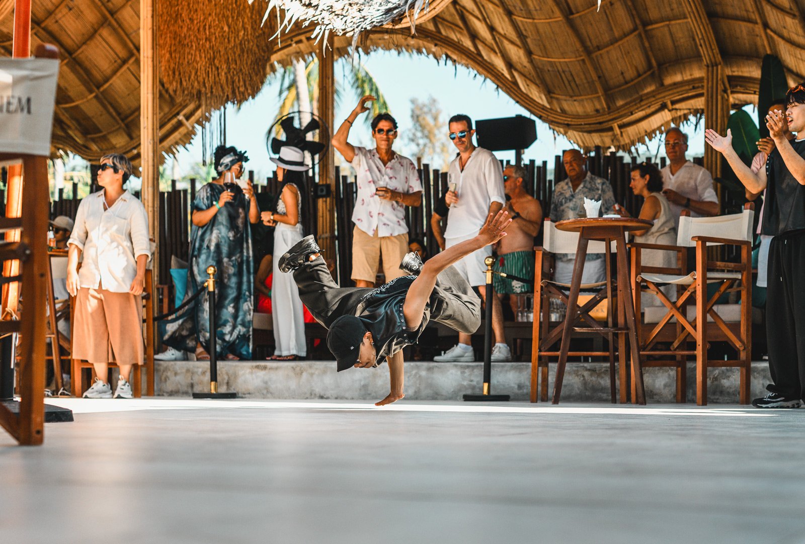 A man breakdancing on a dance floor at a party with a group of people watching and clapping in a tropical outdoor venue.