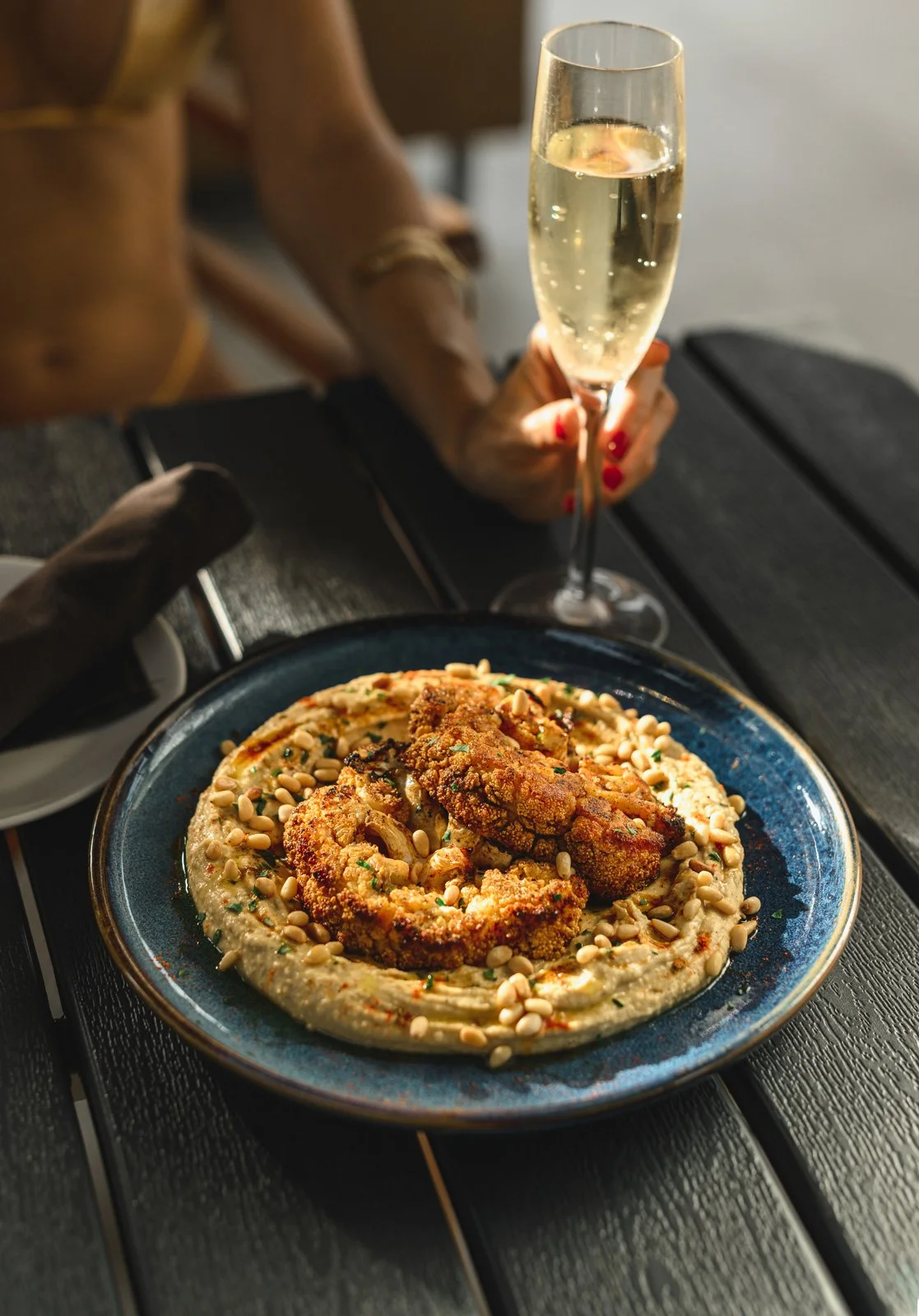 A plate of fried chicken served with a side of hummus topped with pine nuts, garnished with herbs, on a dark blue plate. In the background, a person with red nail polish is holding a glass of sparkling wine.
