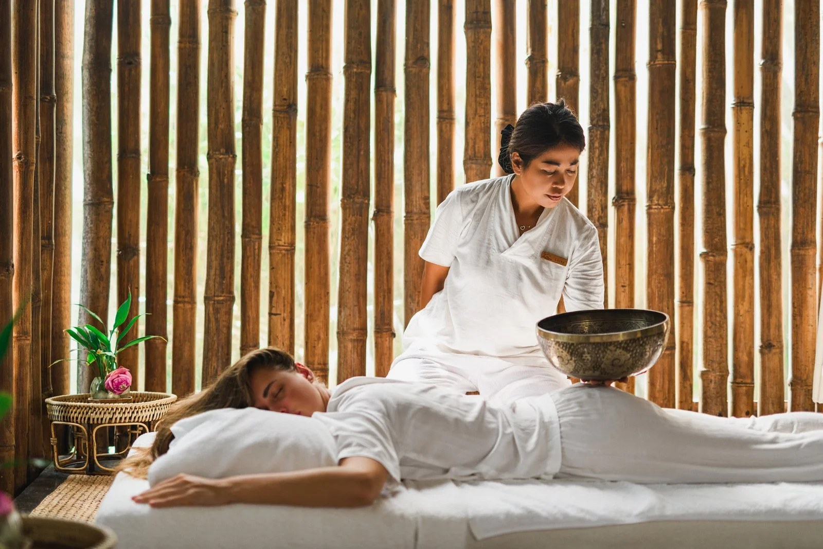 Woman receiving a massage in a spa, lying face down on a massage table, with a massage therapist applying oil on her back in a bamboo-themed room.
