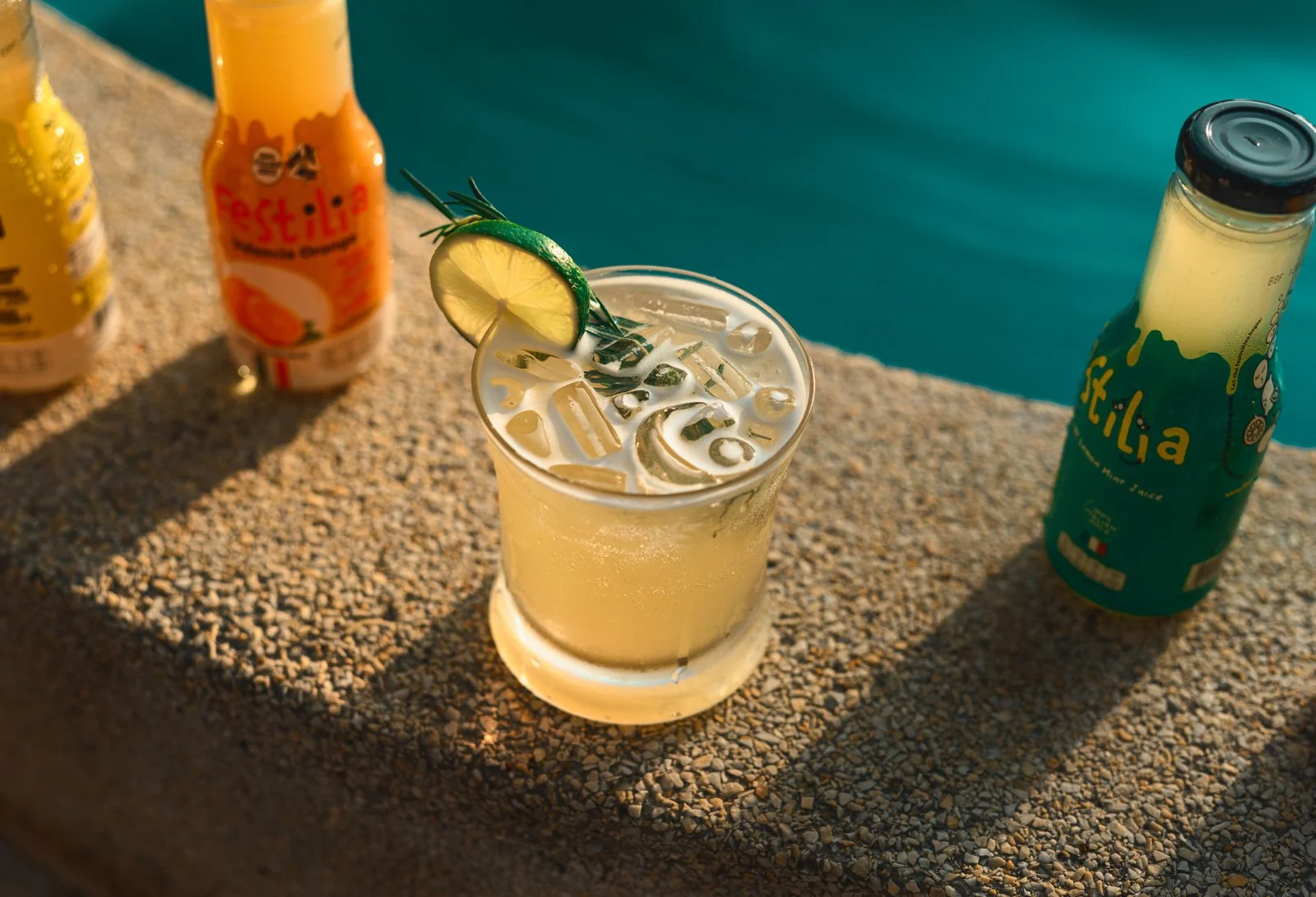 A glass of lemon-flavored beverage with ice and a lemon wedge garnished with a sprig of rosemary, placed on a textured surface near a swimming pool. Two bottles of Stella juice are also on the surface.