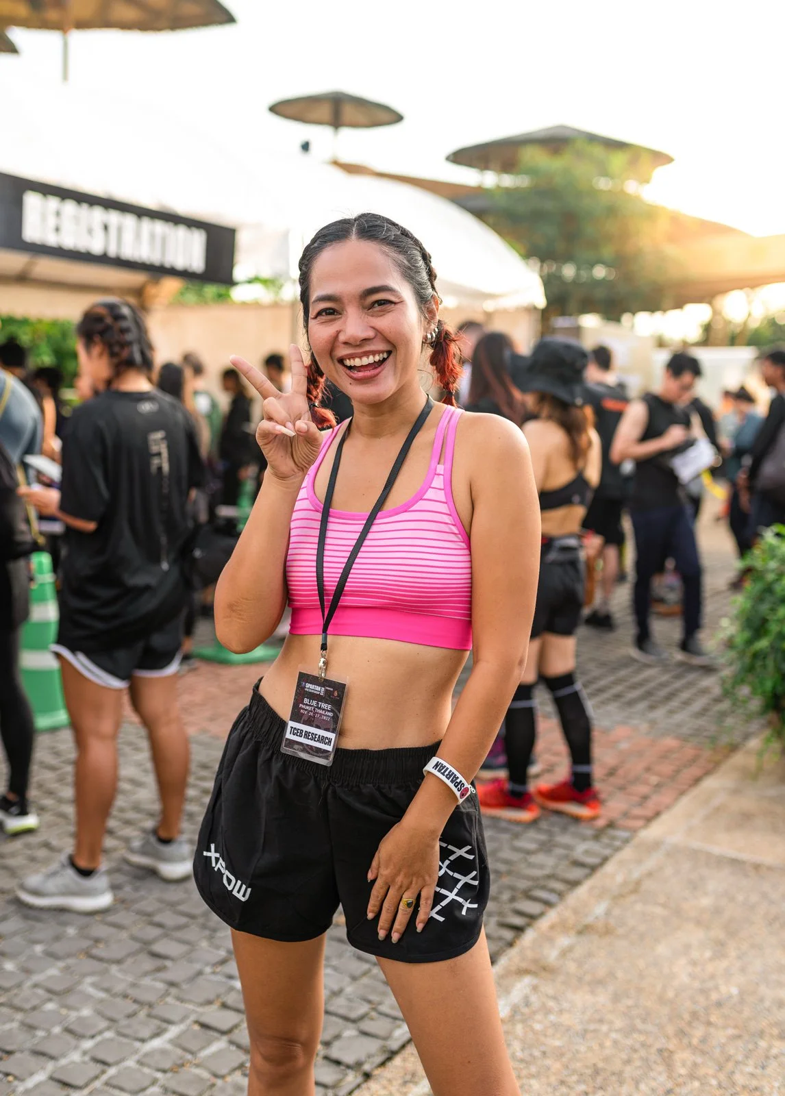 A woman with braided hair, wearing a pink and purple striped sports bra and black shorts, smiling, making a peace sign, and standing outdoors at an event with a crowd in the background.