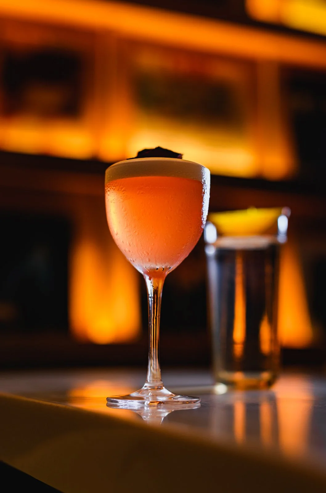 A close-up of a pink cocktail in a coupe glass with a frothy top, condensation on the glass, and a dark, warm-toned background with blurred lights.