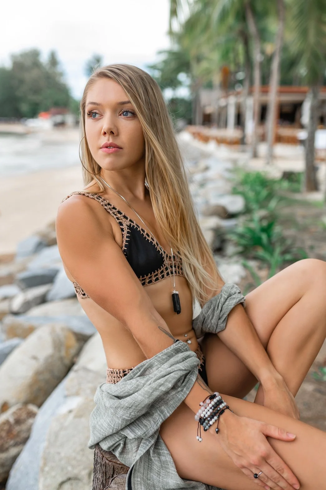 A young woman with long blonde hair sitting outdoors near the beach, wearing a black and beige crochet bikini top and jewelry, looking to the side with a calm expression.