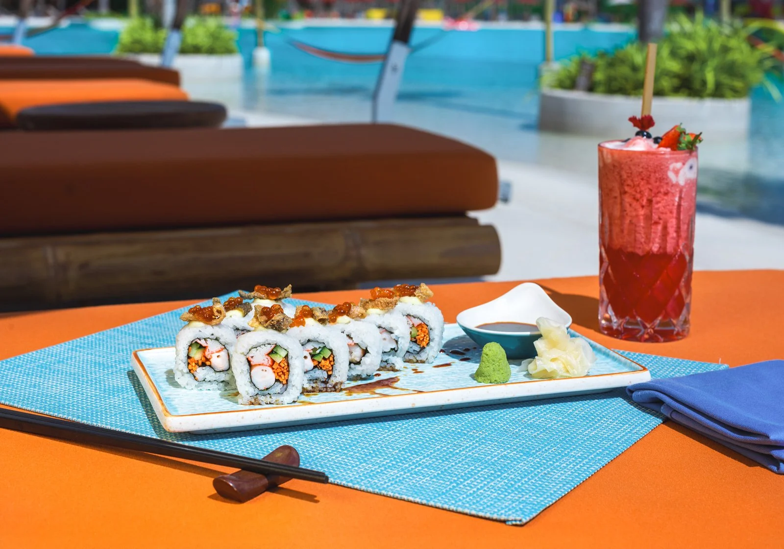 A plate of sushi rolls with garnishes, a small dish of soy sauce, wasabi, and pickled ginger on a table with a blue placemat, a blue napkin, and chopsticks, with a pink tropical cocktail decorated with fruit and berries in the background by a pool.