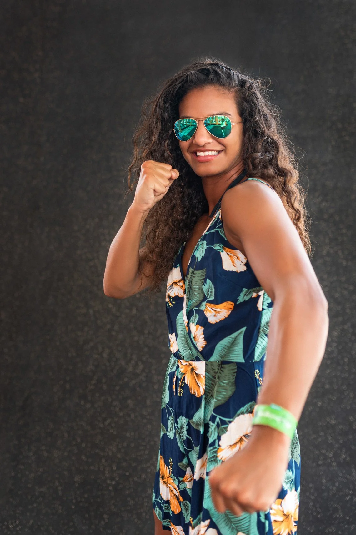 A woman with curly hair wearing reflective sunglasses and a floral dress, smiling and making a fist gesture.
