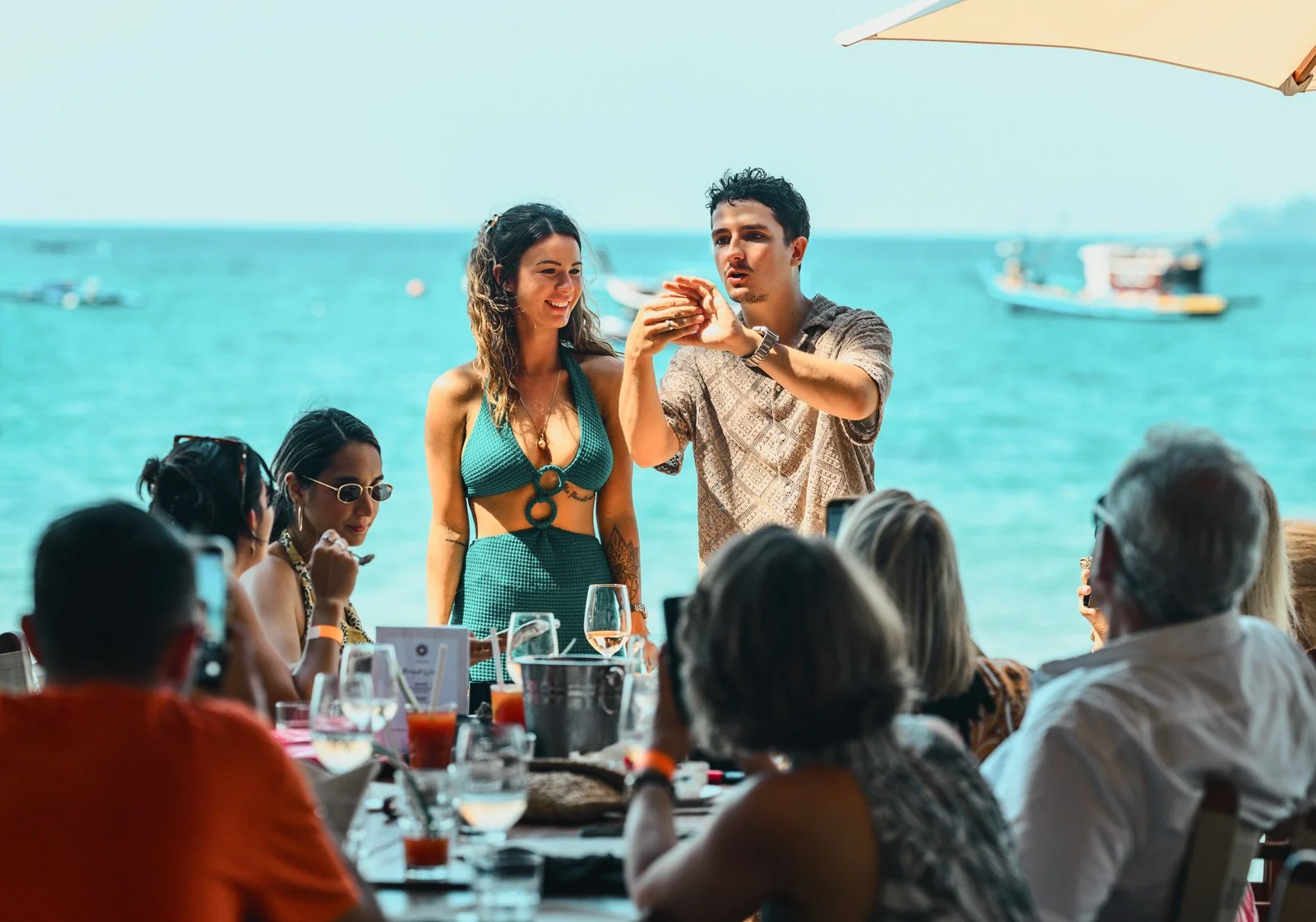 A man and a woman perform a magic trick or demonstration to a group of people seated at a table near the waterfront, with boats on the water and an umbrella overhead.