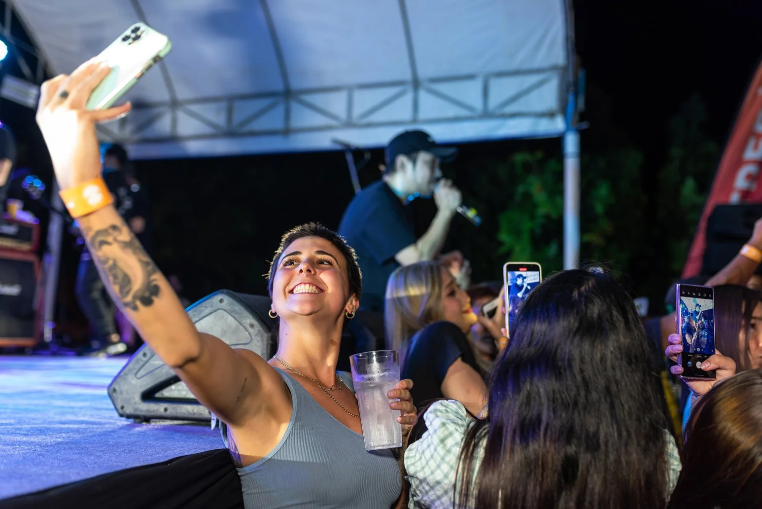 A young woman with short dark hair and tattoos on her arm is smiling and taking a selfie at a concert, holding a drink in her other hand. People around her are also taking photos. A performer on stage is singing into a microphone in the background un