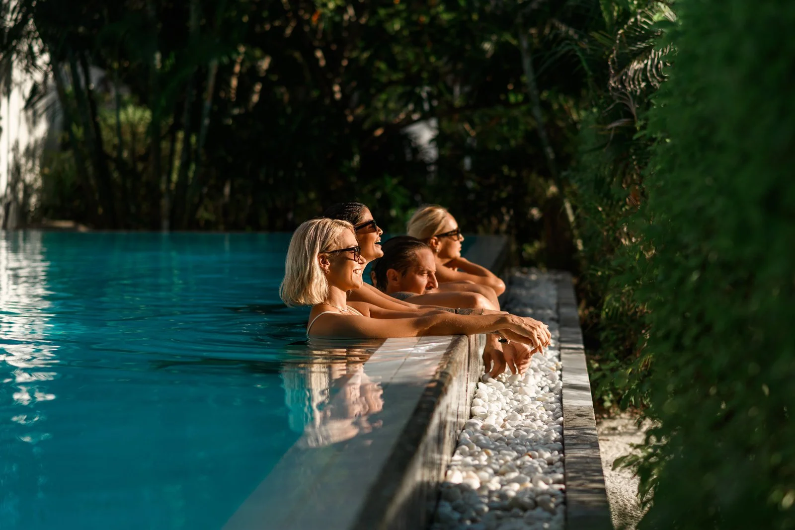 Group of five smiling women in sunglasses leaning on the edge of a swimming pool, overlooking lush green foliage.