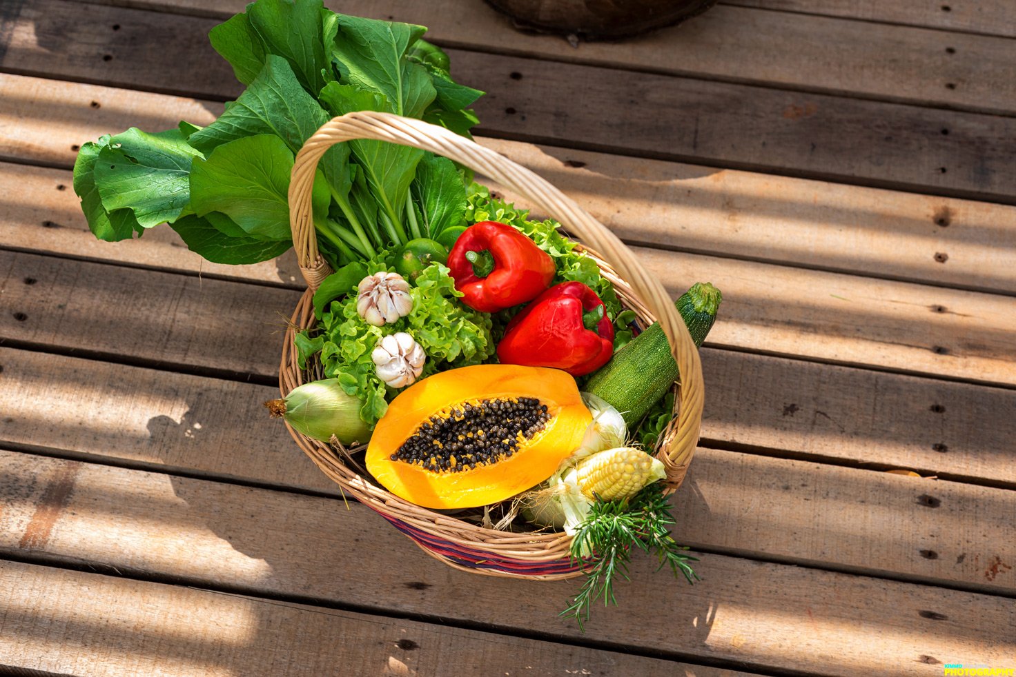 A basket of fresh vegetables including green leafy lettuce, garlic, red bell peppers, zucchini, a halved yellow papaya with black seeds, an onion, a small ear of corn, and sprigs of herbs on a wooden surface.