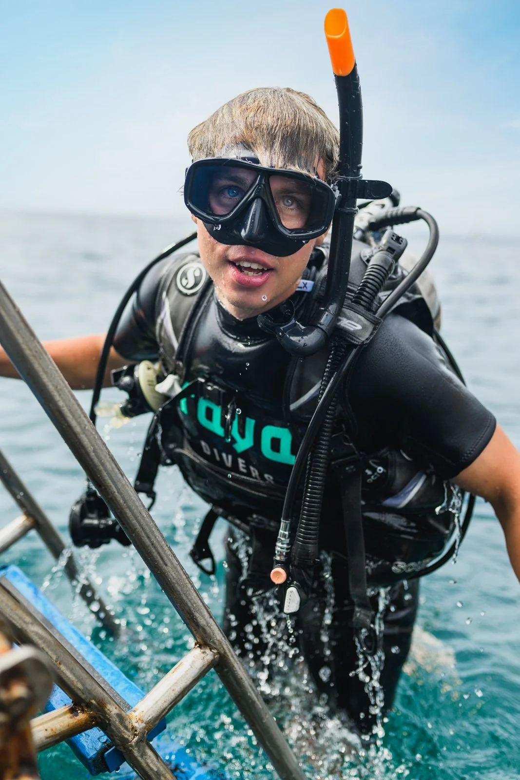 A young man in scuba diving gear, including a mask, snorkel, wetsuit, and tank, climbing out of the water onto a boat ladder with the ocean in the background.
